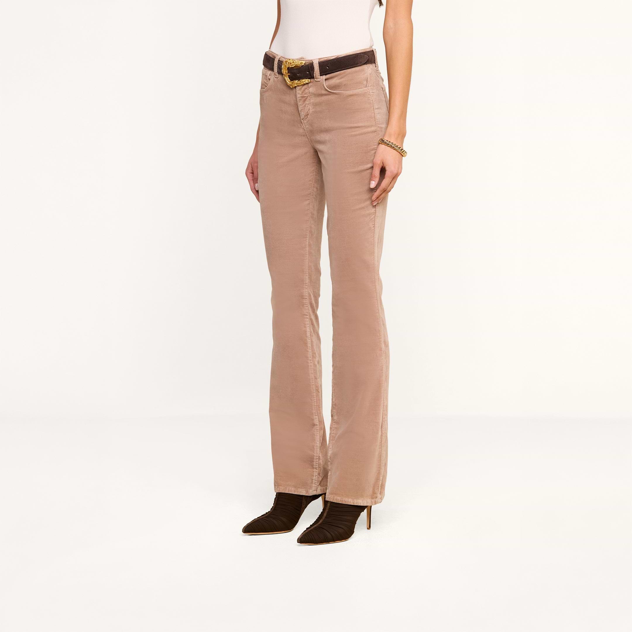 A person wearing the Stevie Velvet Jean in light brown, styled with a white top, a dark brown belt featuring a gold buckle, a bracelet, and dark heeled boots stands against a plain white background.