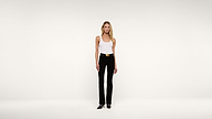 A woman stands against a plain white background, wearing a white tank top, black pants with a belt, and black shoes.