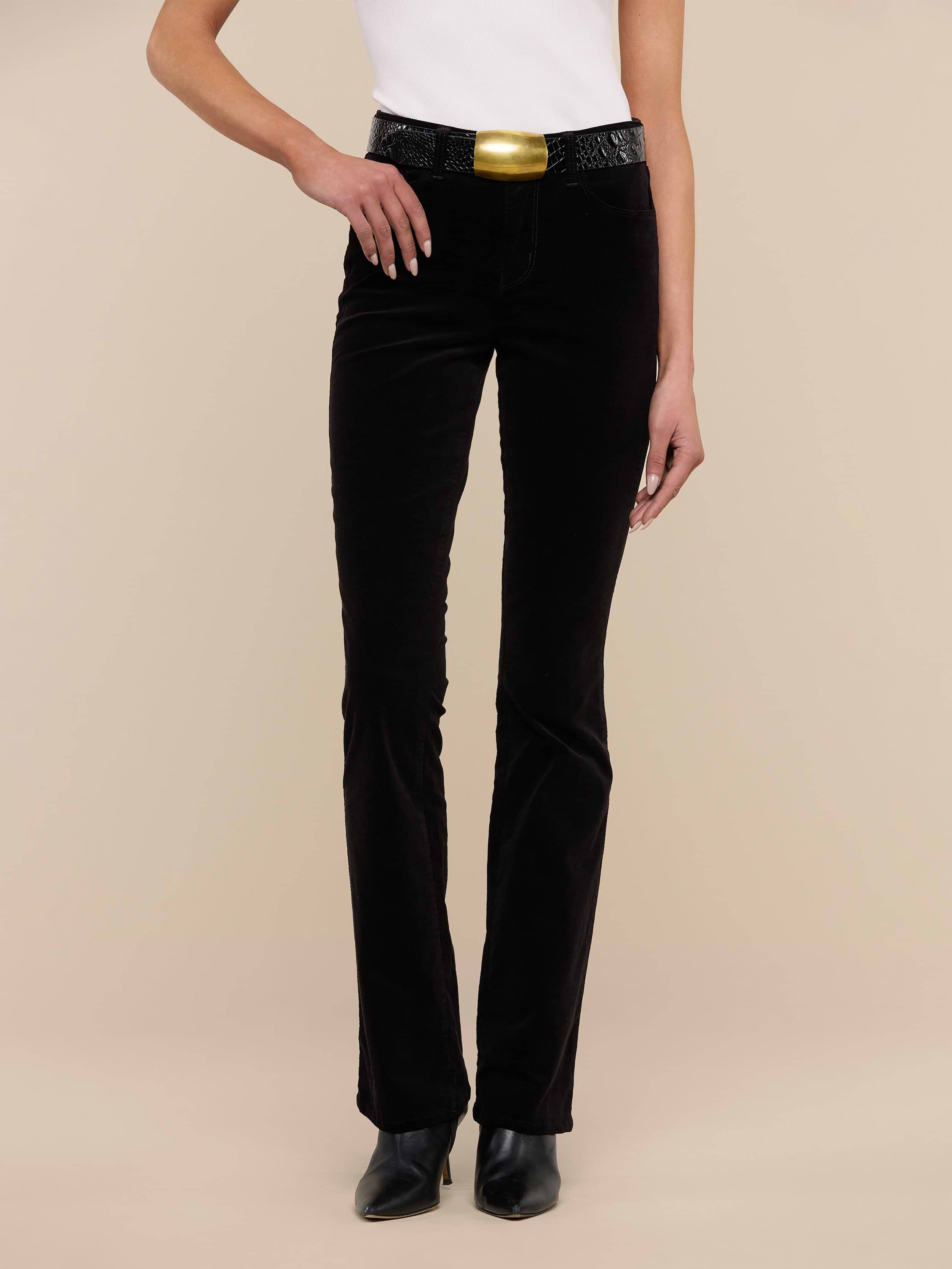 A person wearing a white sleeveless top, the Stevie Velvet Jean in black, black shoes, and a large gold belt buckle stands against a plain white background.