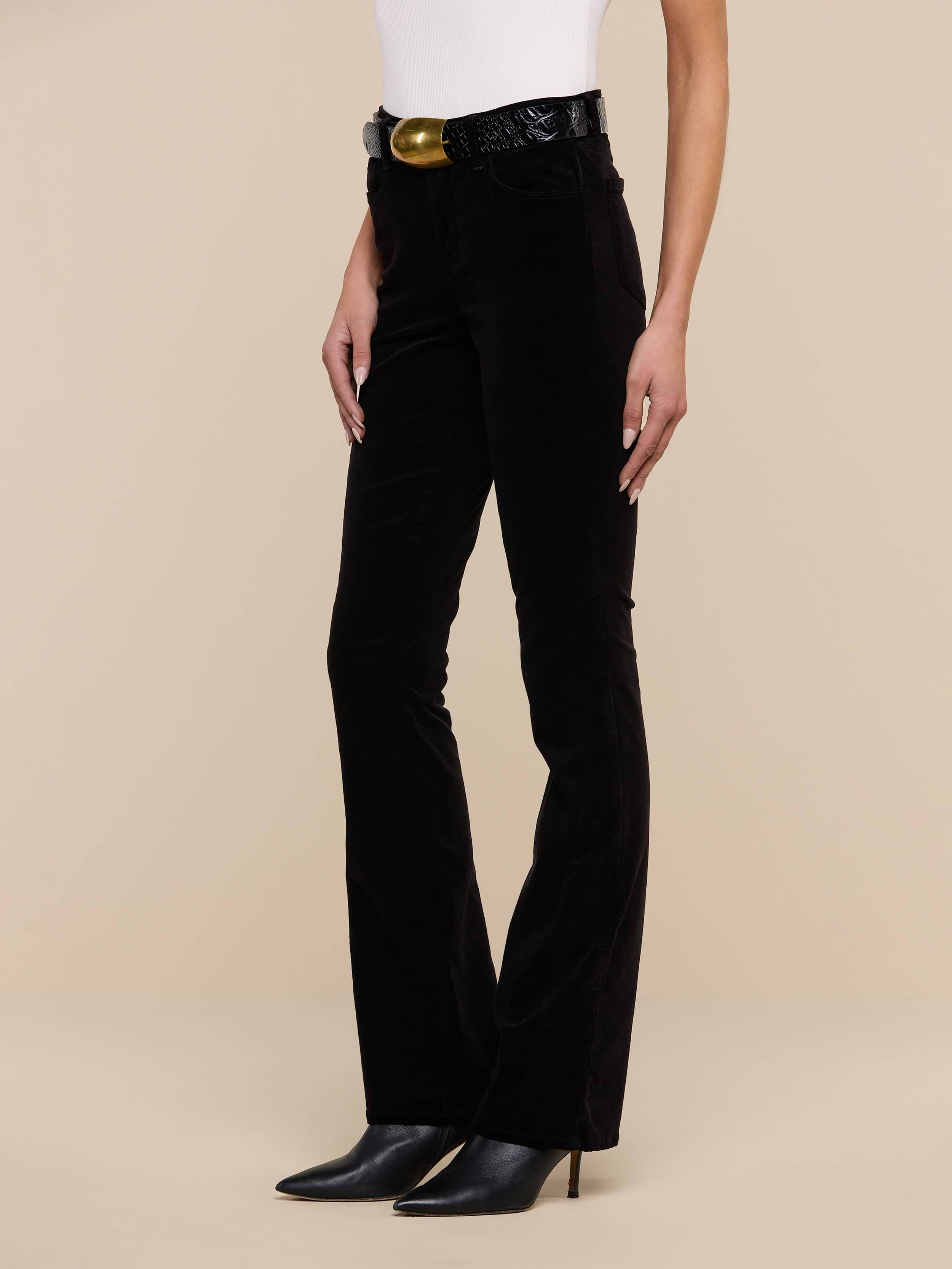 A person wearing the Stevie Velvet Jean in black, paired with a white sleeveless top, a black belt with a gold buckle, and black pointed-toe heeled boots stands against a plain white background.