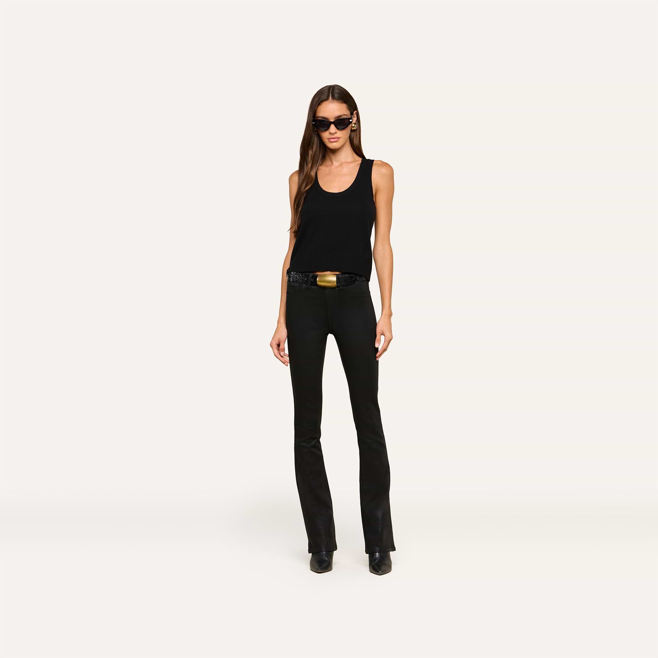 A woman stands against a plain light background, facing forward with her hands by her sides, wearing sunglasses, a sleeveless black top, black boots, and the Selma Feather Coated Jean.