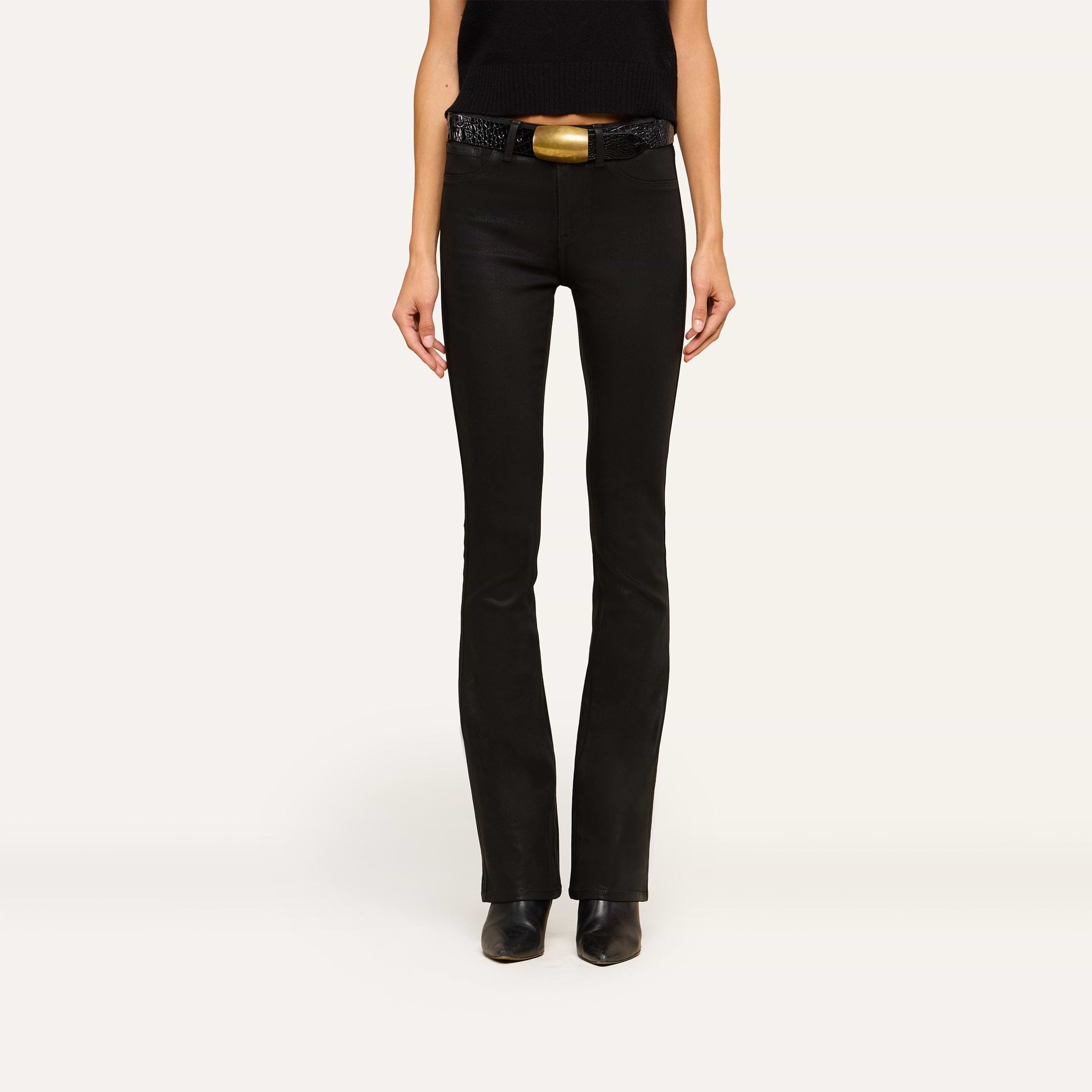 A person stands facing forward wearing the Selma Feather Coated Jean in black, styled with a black top, a black belt featuring a large gold buckle, and black pointed-toe boots against a plain light background.