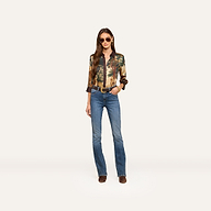 A woman stands confidently against a plain light background, wearing sunglasses, a patterned long-sleeve shirt, a belt, brown boots, and the Selma Bootcut Jean.