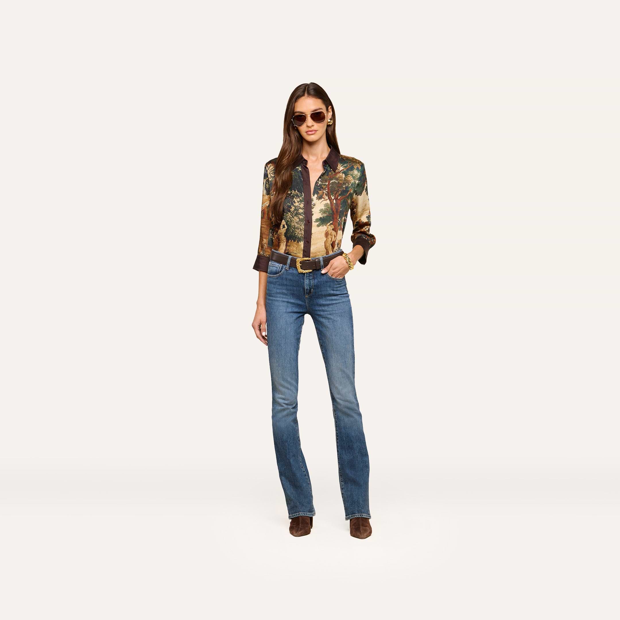 A woman stands confidently against a plain light background, wearing sunglasses, a patterned long-sleeve shirt, a belt, brown boots, and the Selma Bootcut Jean.