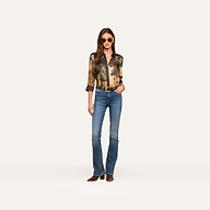 A woman wearing sunglasses, a patterned blouse, the Selma Bootcut Jean crafted from stretch denim, and brown boots stands confidently against a plain light background.