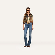 A woman stands against a plain white background wearing sunglasses, a printed long-sleeve shirt, a brown belt, brown shoes, and the Selma Bootcut Jean made from stretch denim.
