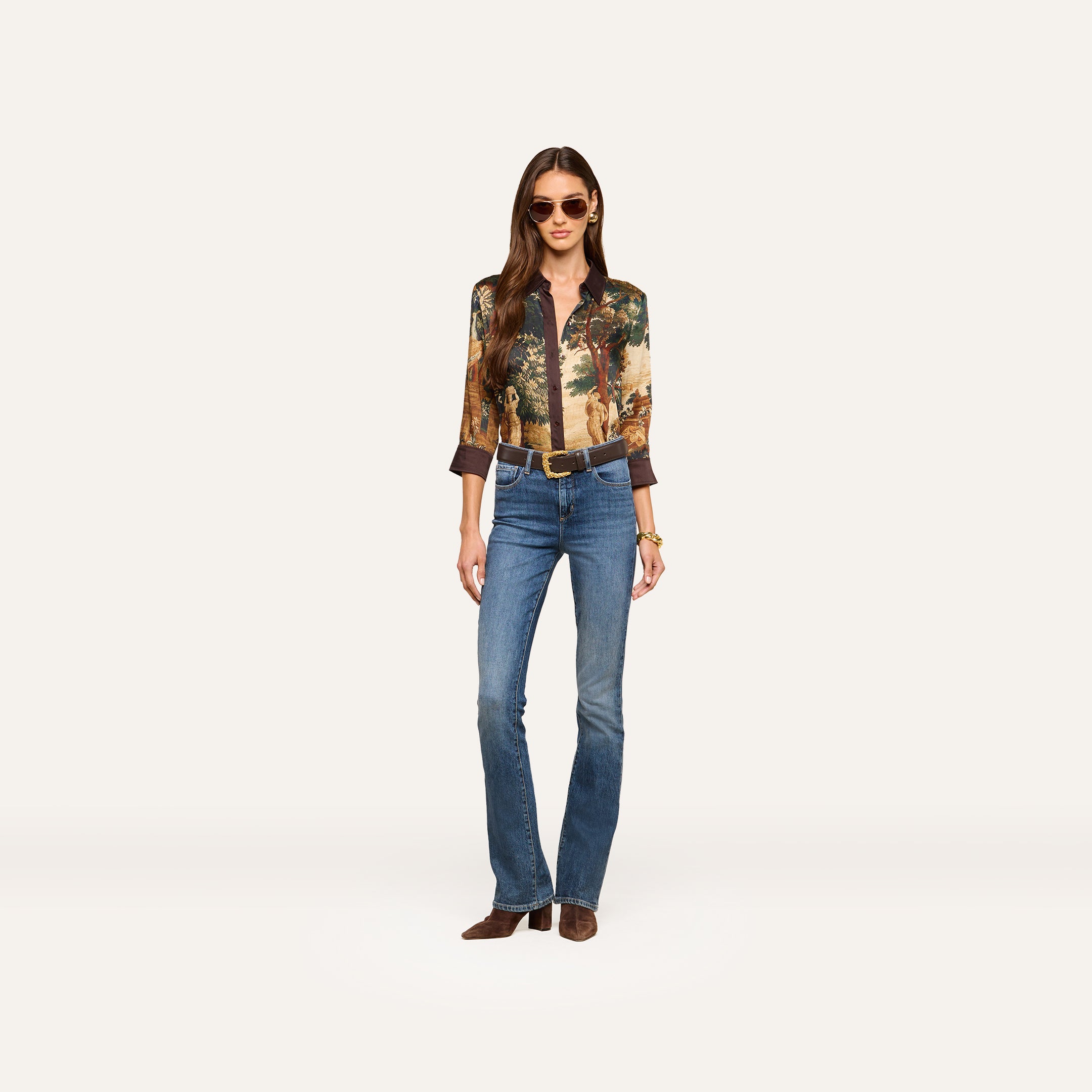 A woman stands against a plain white background wearing sunglasses, a printed long-sleeve shirt, a brown belt, brown shoes, and the Selma Bootcut Jean made from stretch denim.