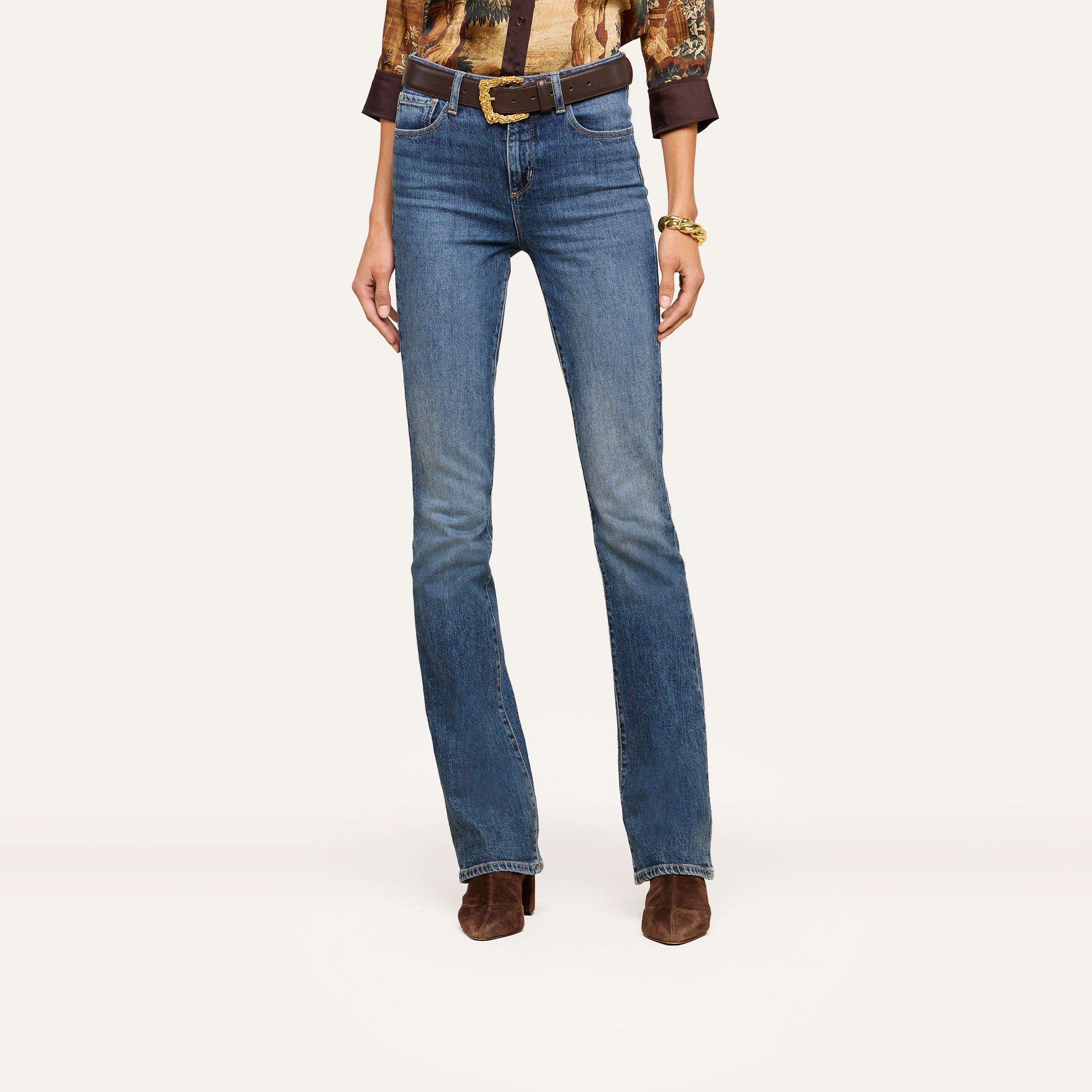A person wearing blue bootcut jeans, a patterned blouse, a brown belt with a gold buckle, a gold bracelet, and brown heeled boots stands against a plain white background.
