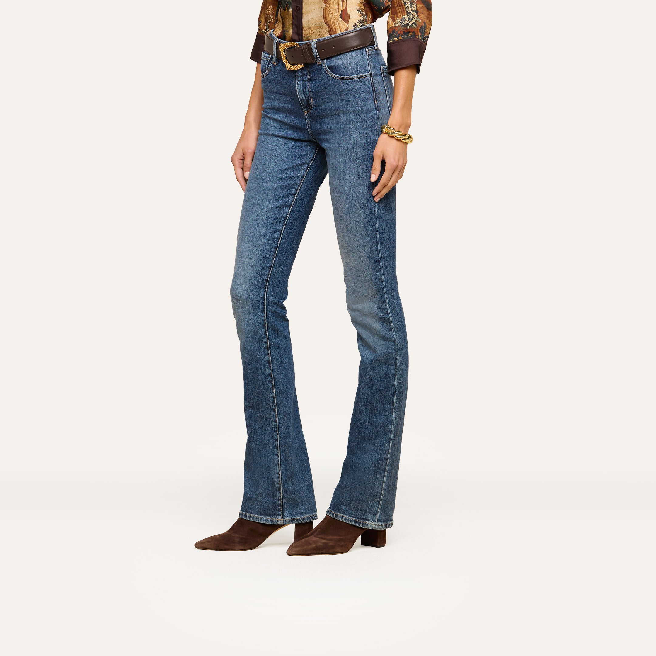Wearing blue bootcut jeans, brown pointed-toe boots, a brown belt, and a long-sleeve patterned shirt, this individual stands against a plain white background. Upper body and face are not visible in the scene.