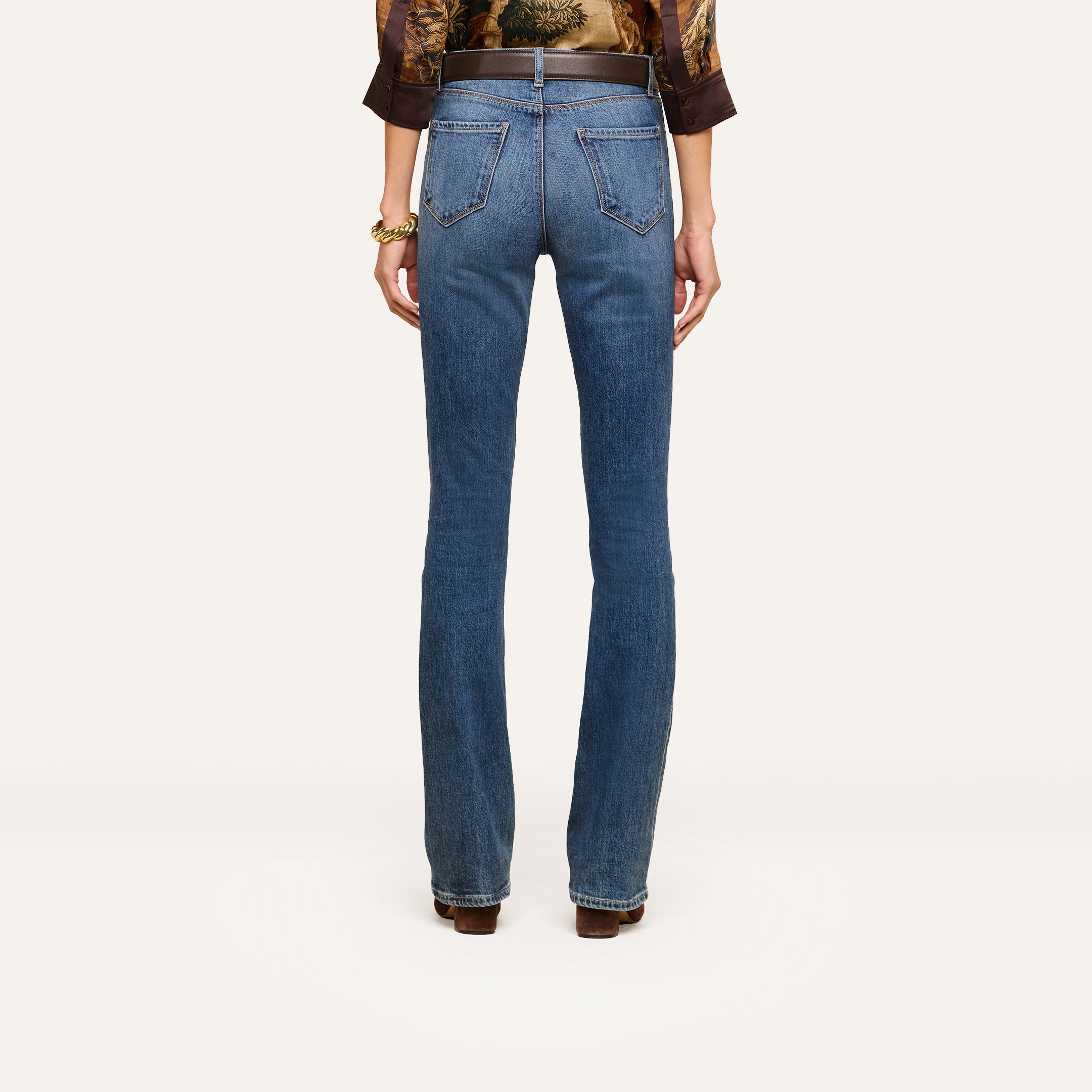 A person stands facing away, wearing the Selma Bootcut Jean—high-rise, blue bootcut jeans crafted from stretch denim with back pockets—and a patterned top. They accessorize with brown shoes and a bracelet, against a plain light-colored background.