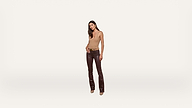 A woman stands against a plain white background wearing a beige sleeveless top, brown leather pants, a belt, and tan heels. She has long dark hair and poses with one hand relaxed by her side.