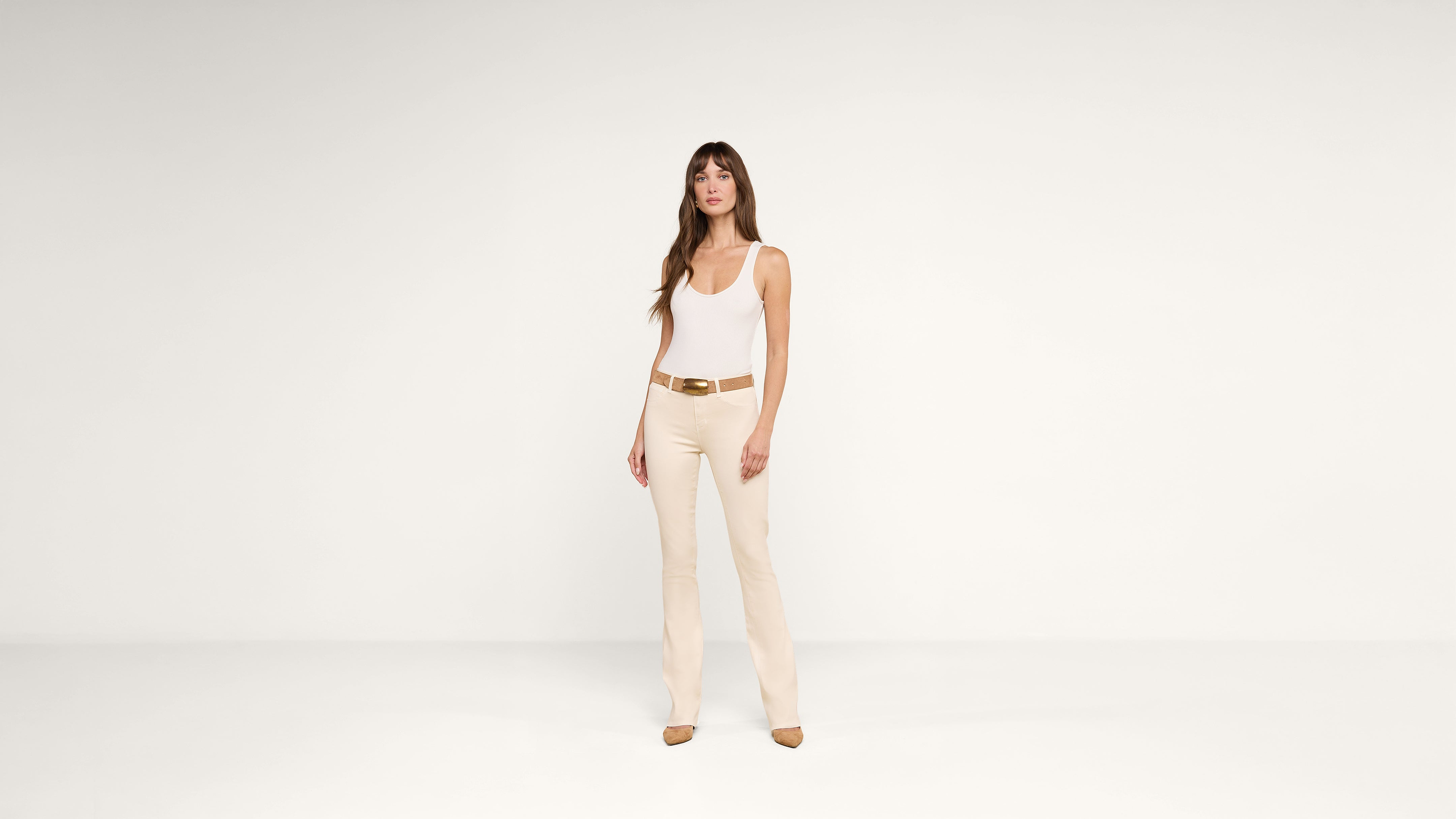 Woman standing against a plain white background, wearing a white tank top, beige pants, a brown belt, and brown heeled shoes. She has long brown hair and a neutral expression.