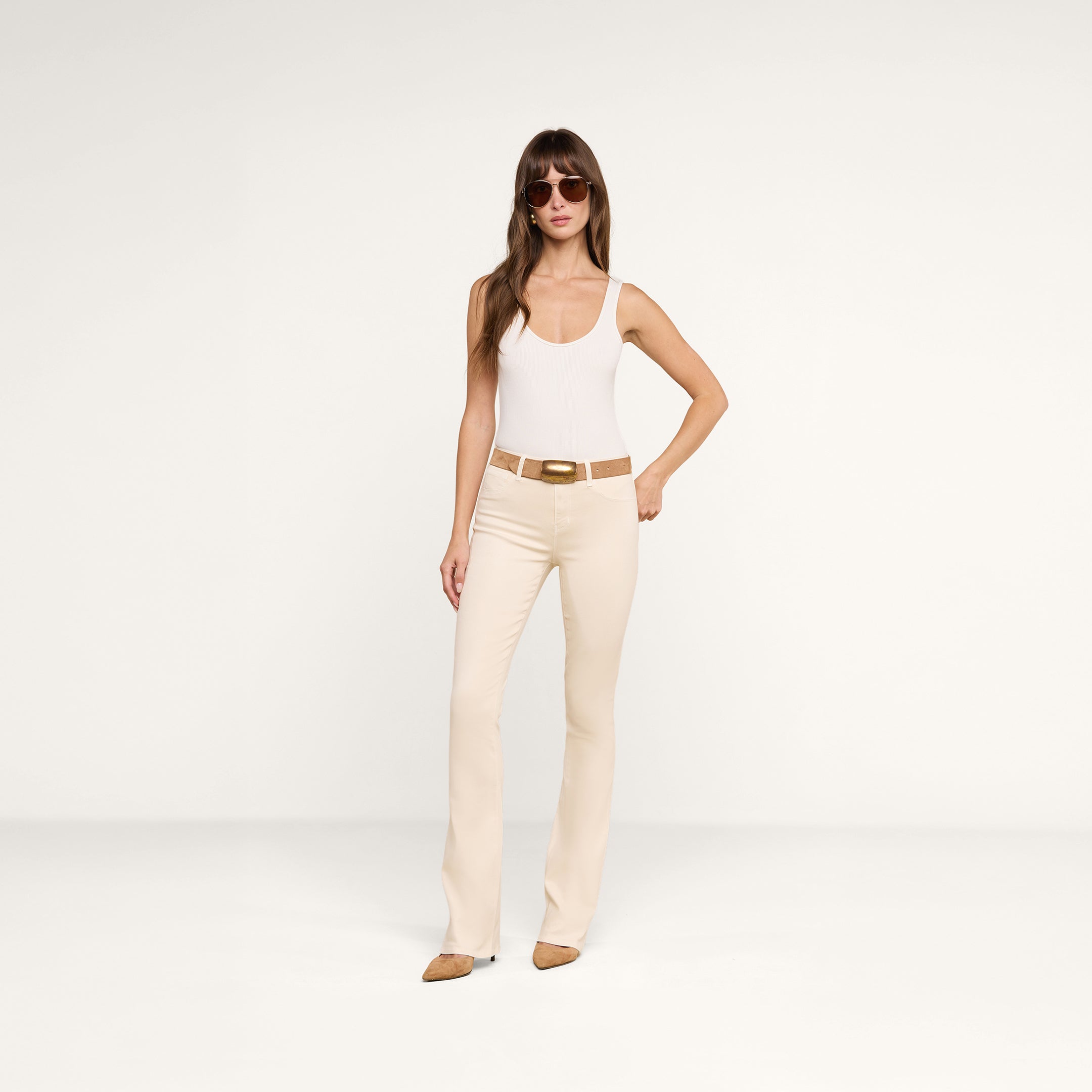 A woman wearing sunglasses, a white tank top, and brown shoes stands confidently against a plain white background, pairing her outfit with the Selma Coated Bootcut Jean.