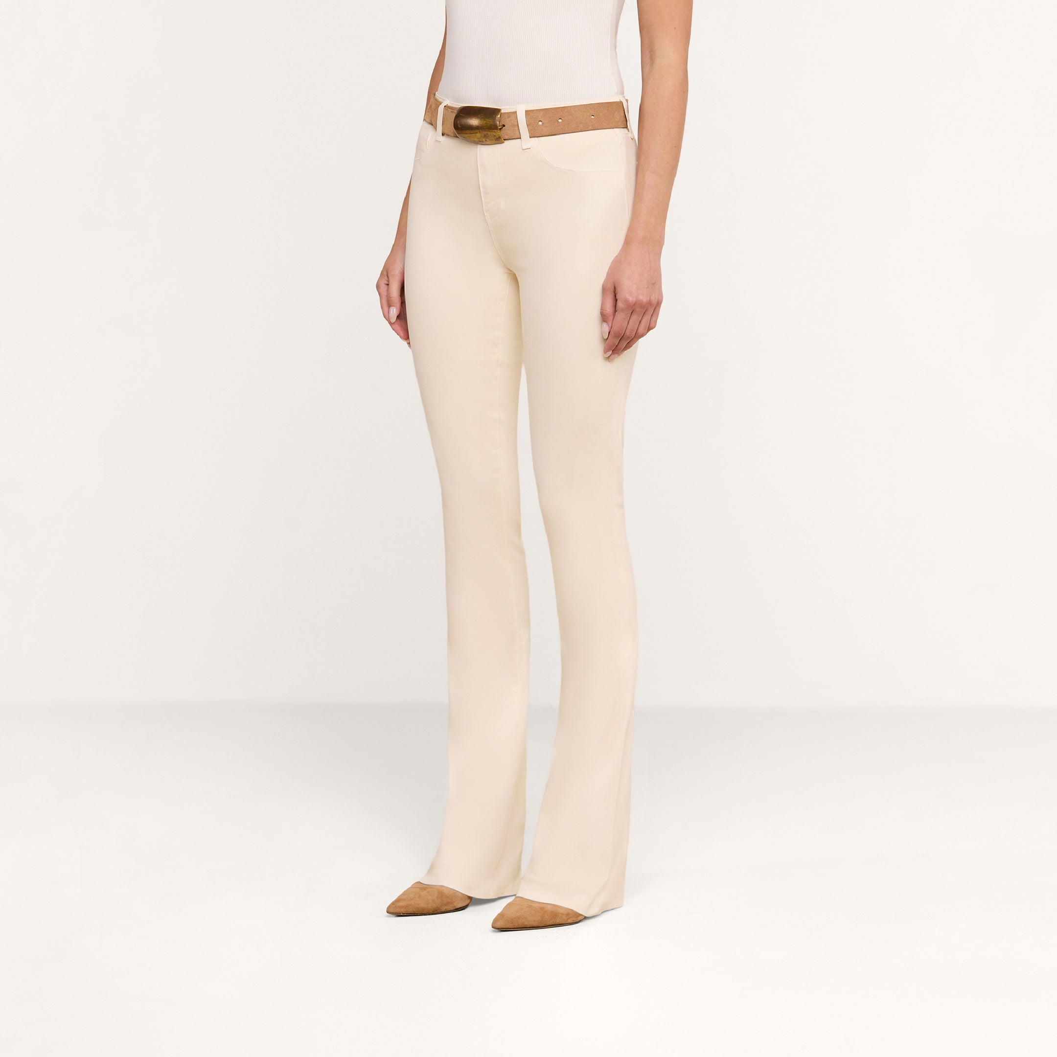 A person wearing the Selma Coated Bootcut Jean in beige, styled with a brown belt, a white top, and brown pointed shoes. They are standing against a plain white background, with their face not visible in the image.