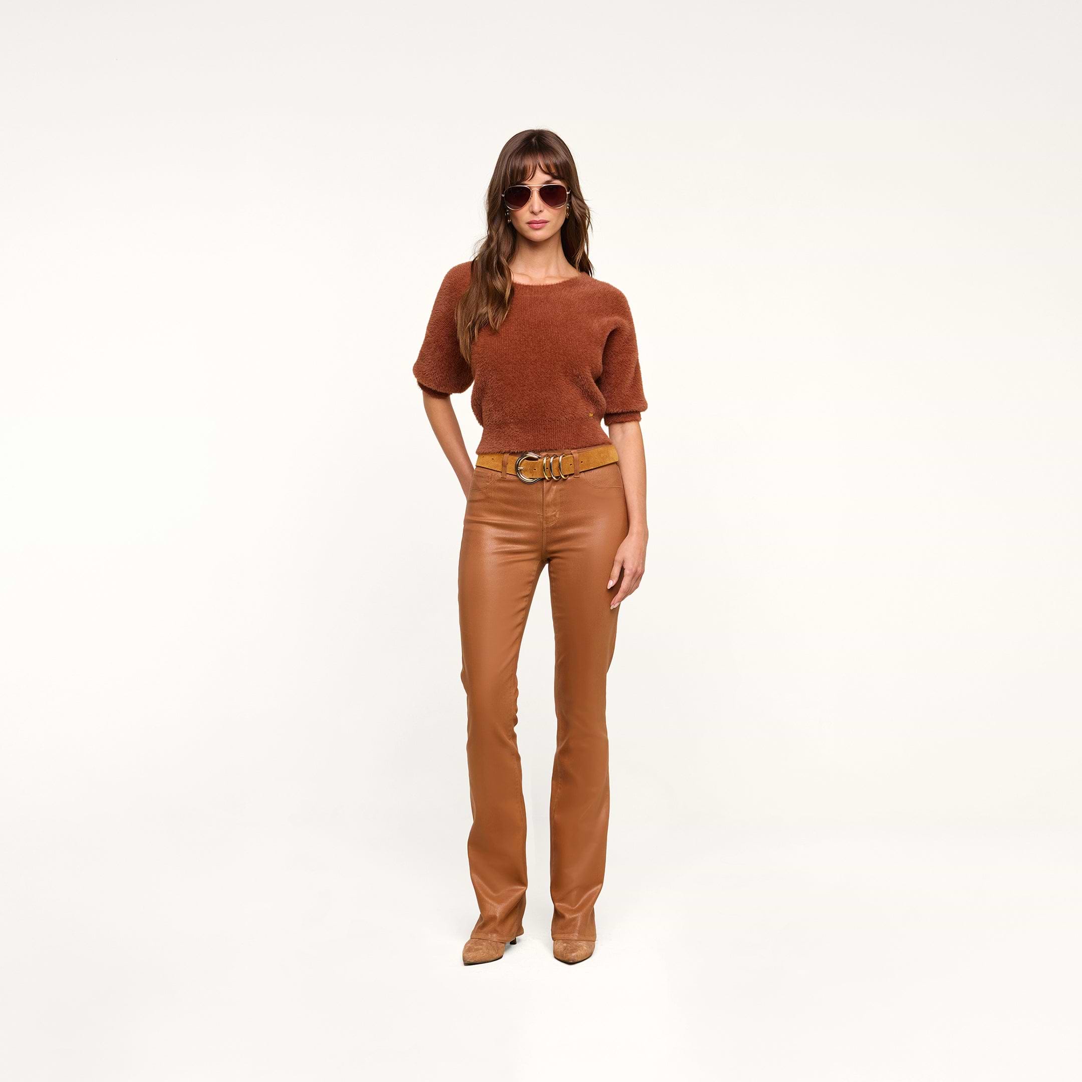 A woman wearing sunglasses, a dark java brown short-sleeve top, tan boots, and the Selma Coated Bootcut Jean stands confidently against a plain white background.