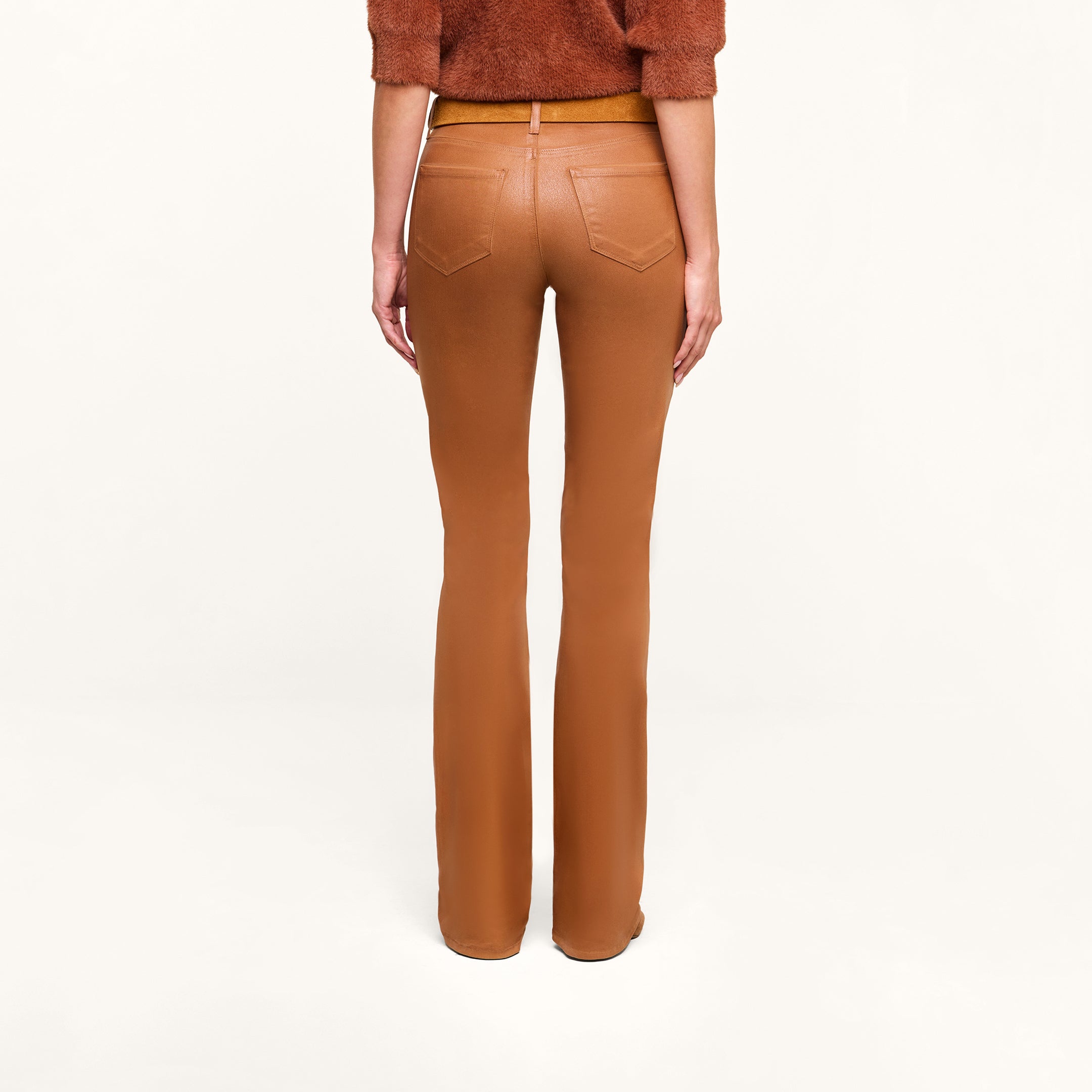 A person models the Selma Coated Bootcut Jean in a high-waisted, camel hue, paired with a dark java brown top, photographed from the back against a plain white background.