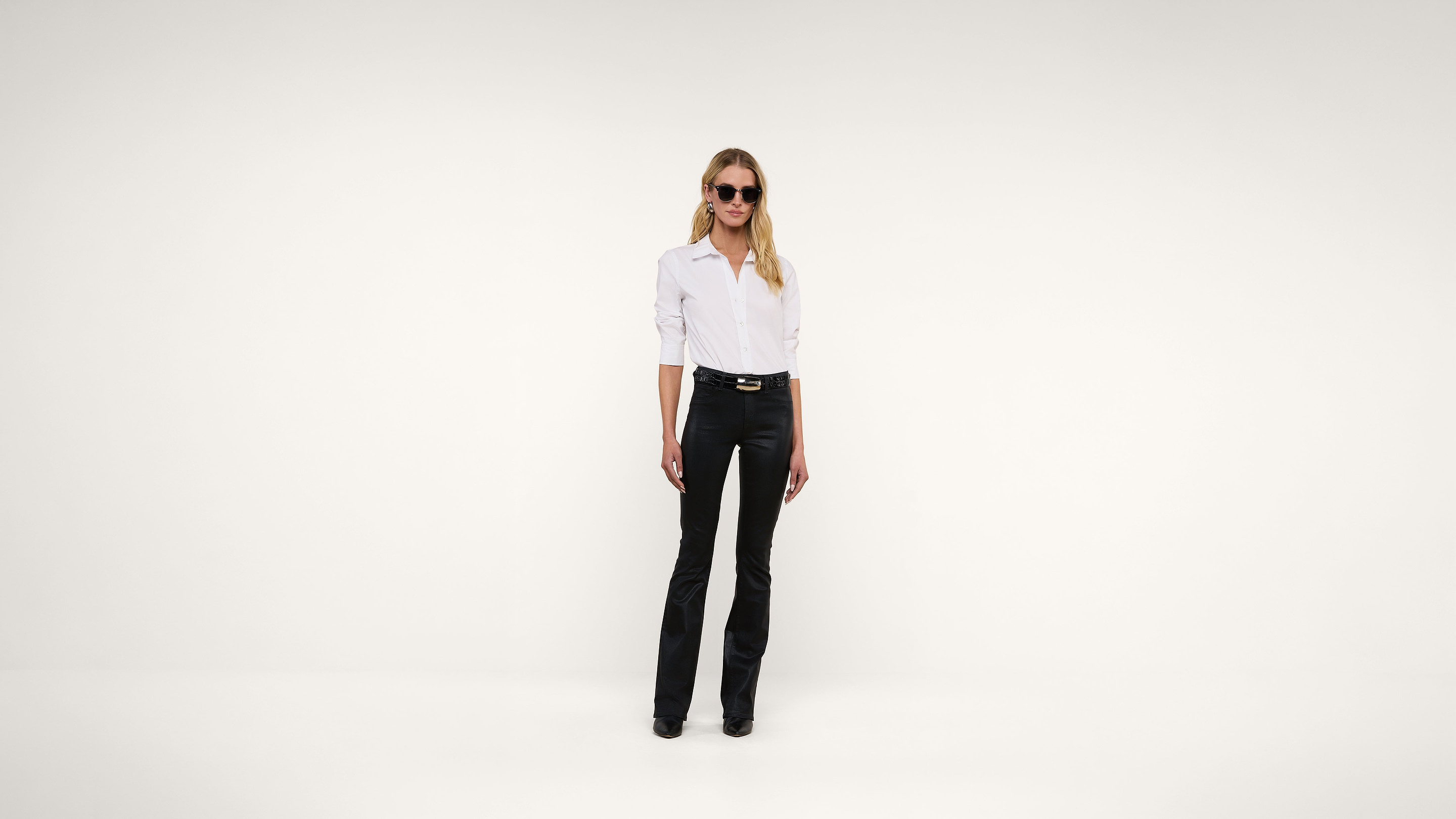 A woman wearing sunglasses, a white button-up shirt, and black pants stands against a plain white background.