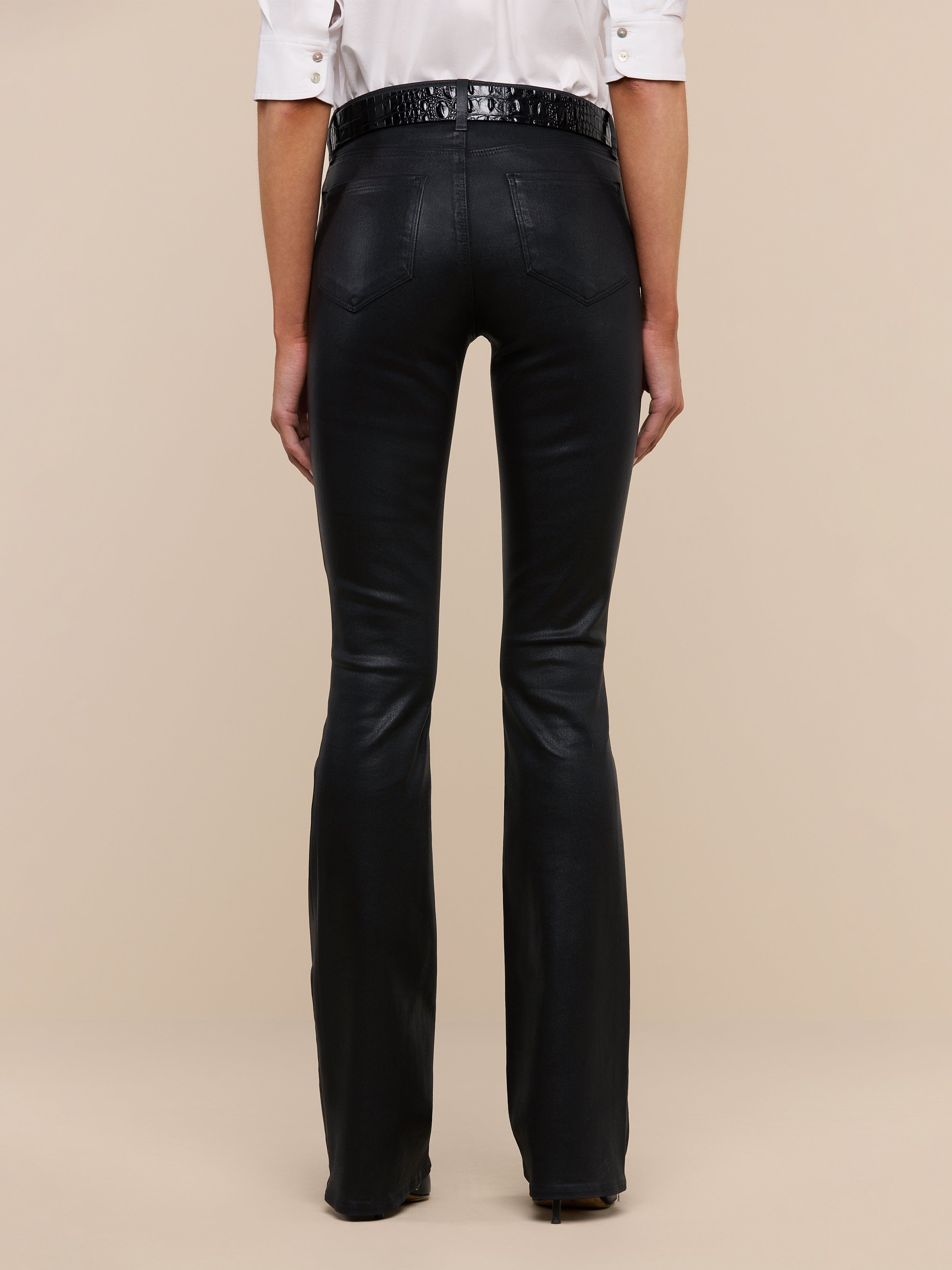 A person stands with their back to the camera, wearing a white shirt and the Selma Coated Bootcut Jean in black. The background is plain and light-colored.
