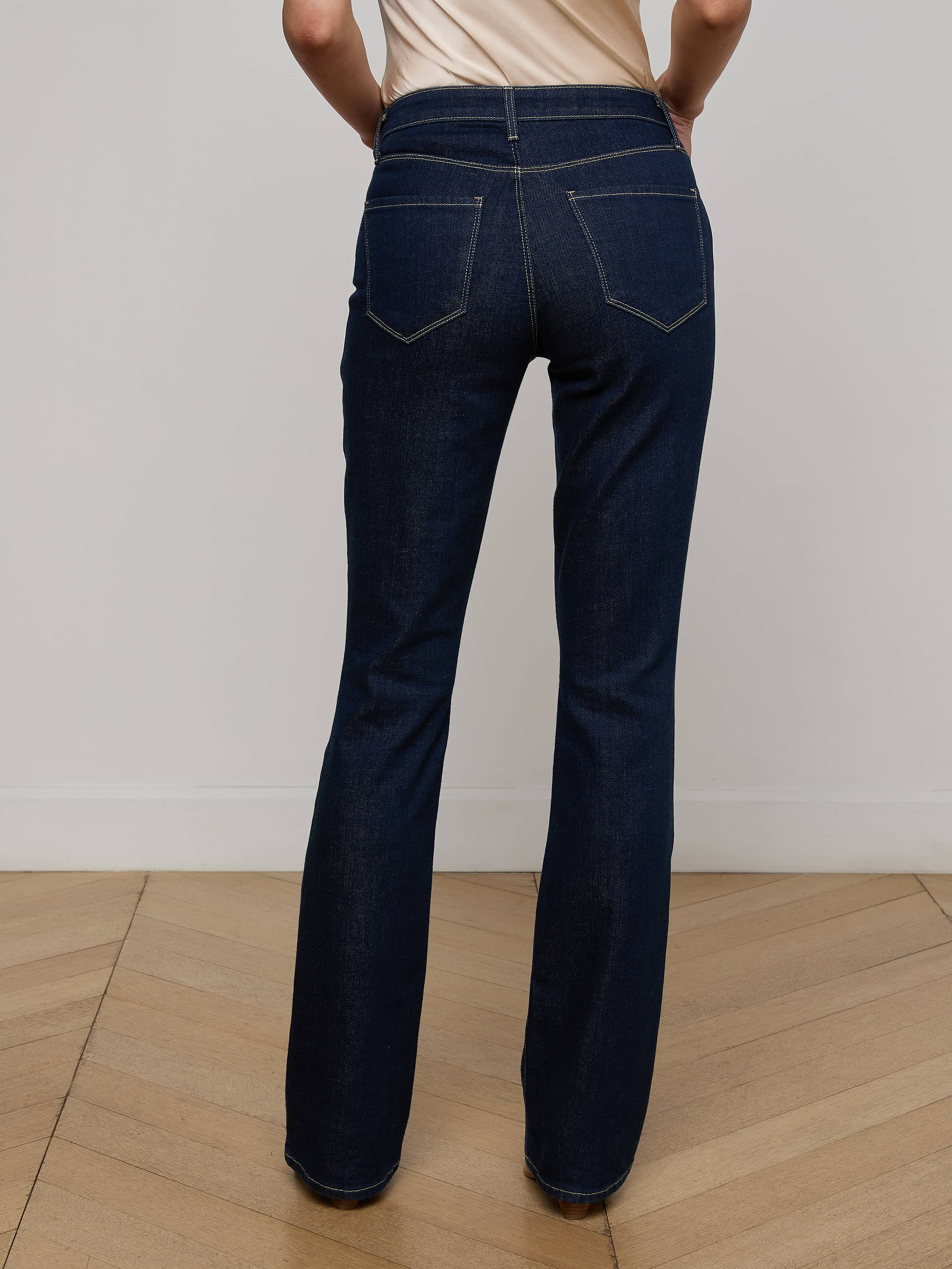 Wearing the Selma Bootcut Jean in dark blue stretch denim paired with a beige top, the person stands with their back to the camera against a plain white background, showcasing the high-rise fit and detailed back pockets of the jeans.