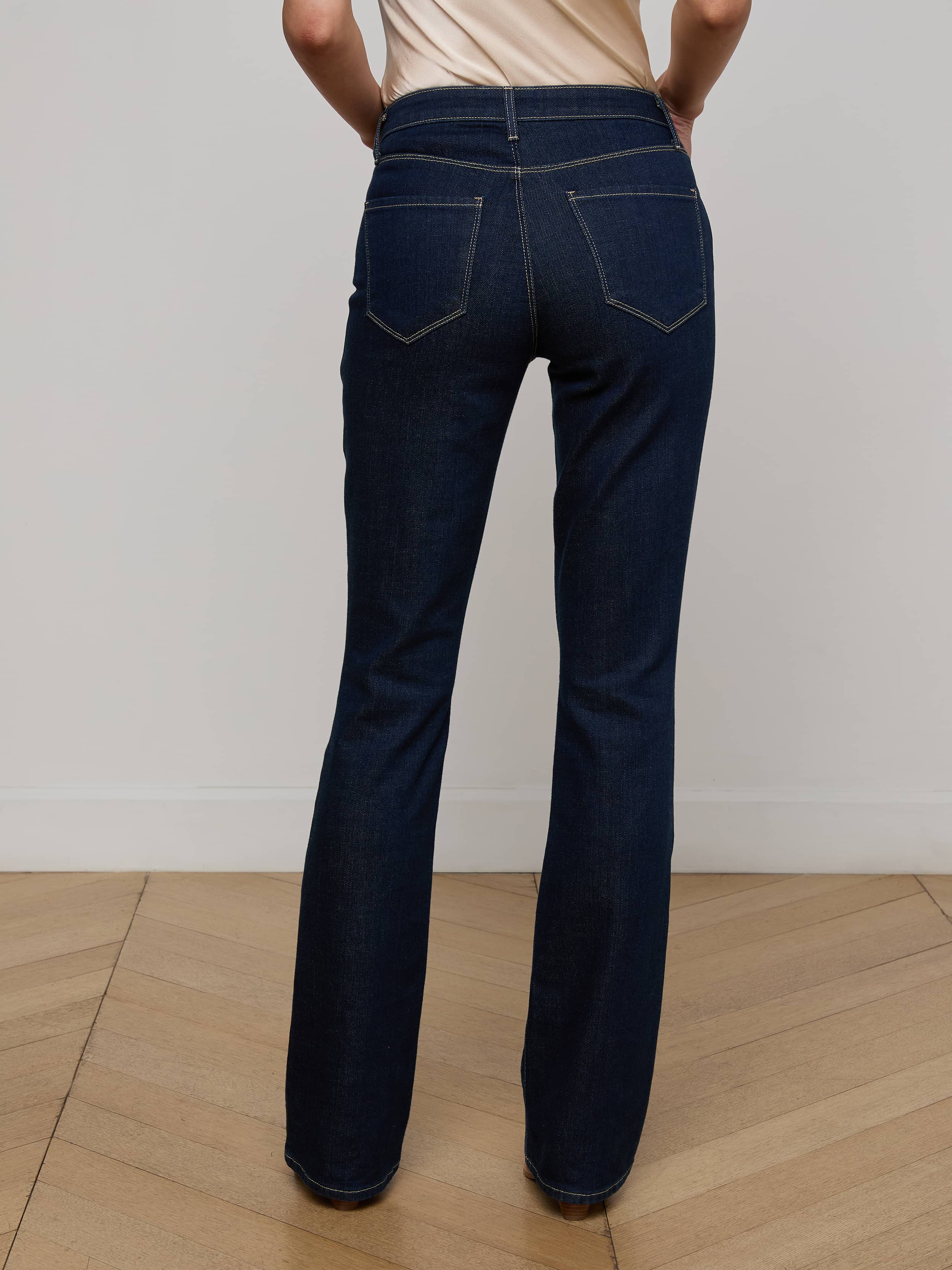 Wearing the Selma Bootcut Jean in dark blue stretch denim paired with a beige top, the person stands with their back to the camera against a plain white background, showcasing the high-rise fit and detailed back pockets of the jeans.