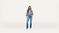 Woman standing against a white background, wearing a sheer long-sleeve blue blouse, high-waisted flare blue jeans, and beige heels, with her left hand in her pocket and her hair styled in loose waves.