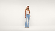 A woman stands against a plain white background wearing a white tank top, light blue flared jeans, a brown belt, and beige shoes. She has straight blonde hair and a neutral expression.