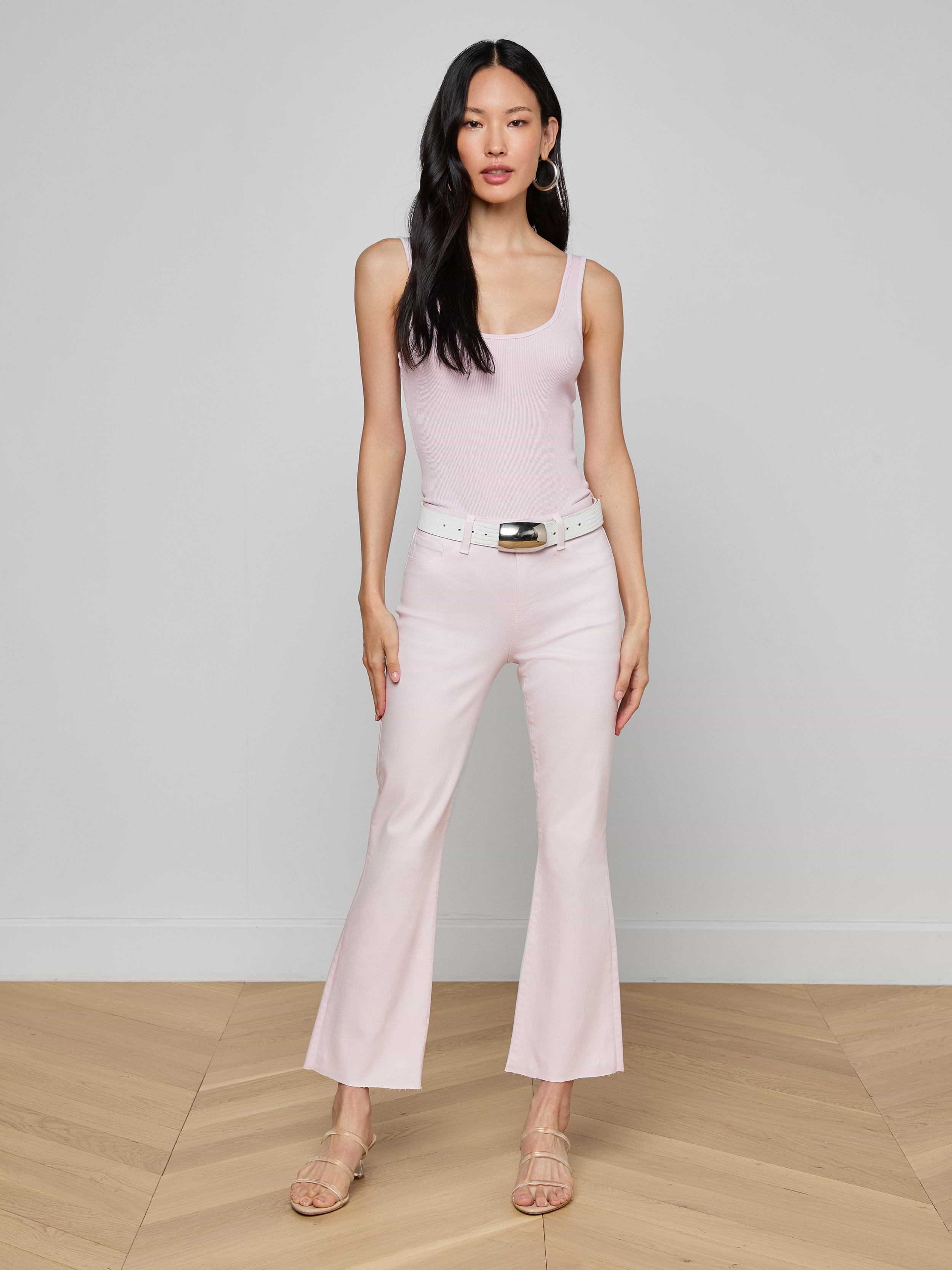 A woman with long dark hair and a neutral expression stands against a plain background, wearing a light pink sleeveless top paired with the Kendra Coated Cropped Flare Jean in a matching shade, styled with a white belt and beige heeled sandals.