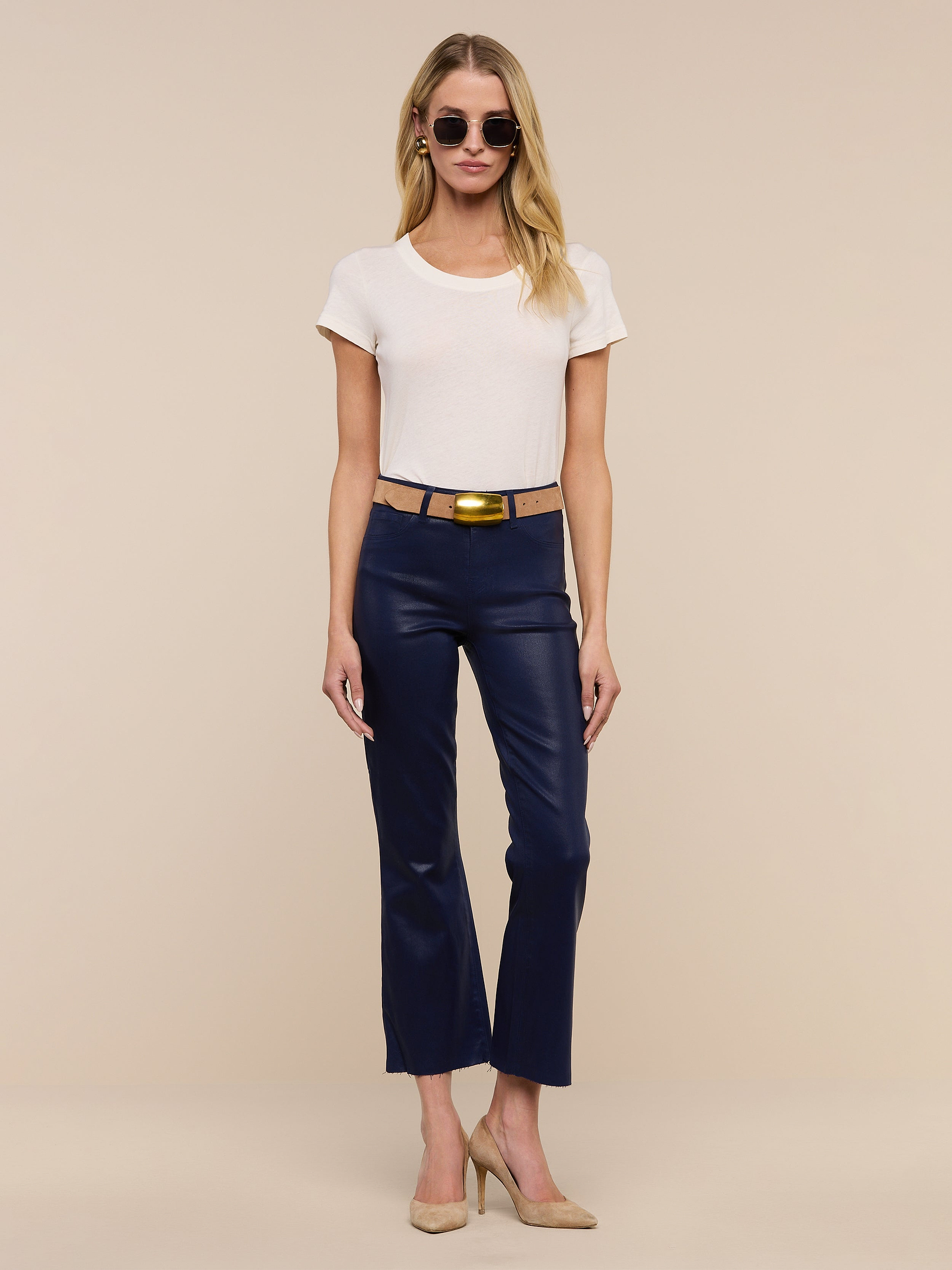 Woman wearing sunglasses, a white t-shirt, and the Kendra Coated Cropped Flare Jean styled with a gold buckle belt and beige heels stands against a plain white background.