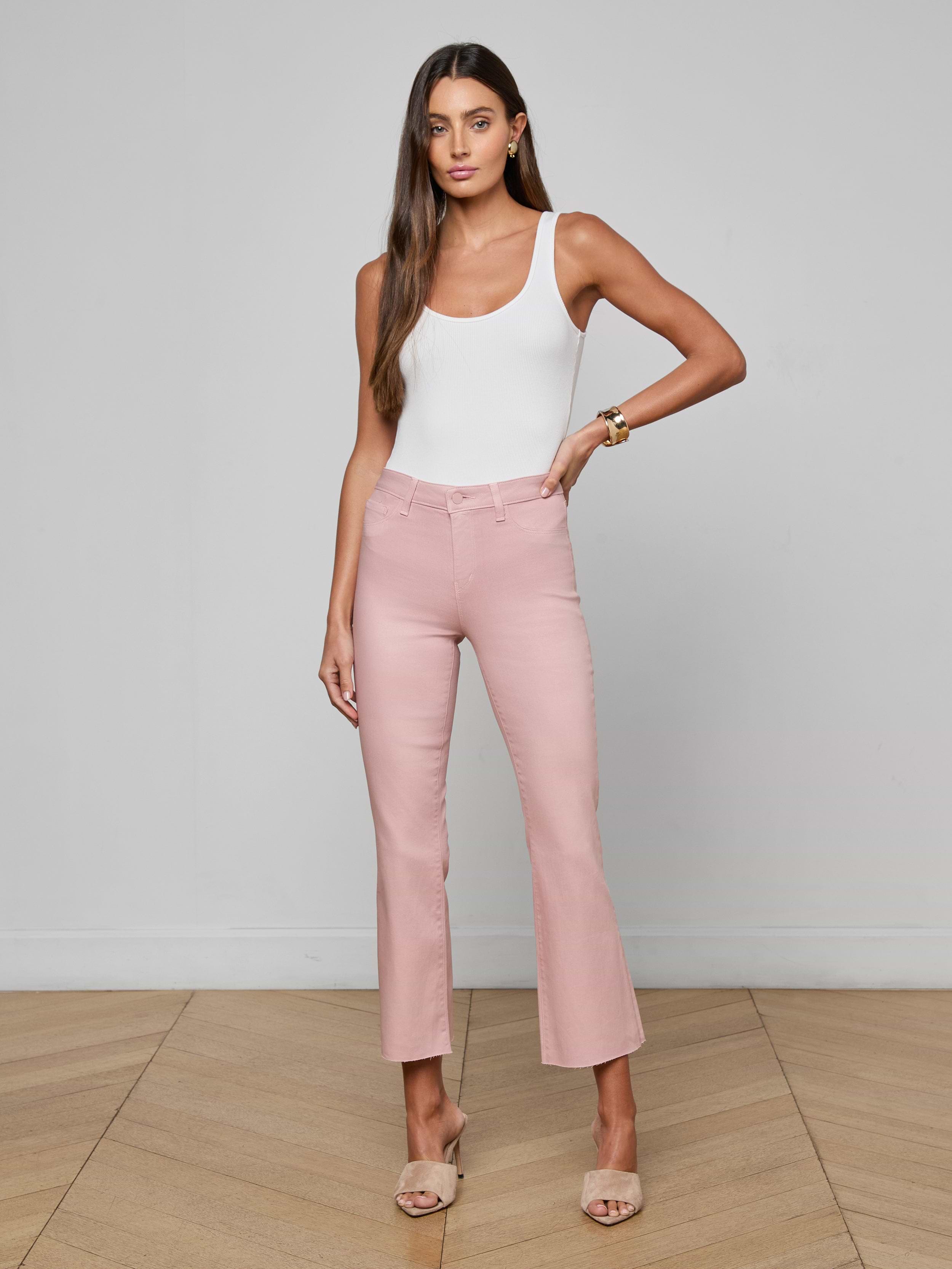 A woman stands against a plain white background wearing a white tank top, the Kendra Coated Cropped Flare Jean in pink—a high-waisted, leather-alternative coated denim—and beige open-toe heels. She has long hair and a neutral expression.