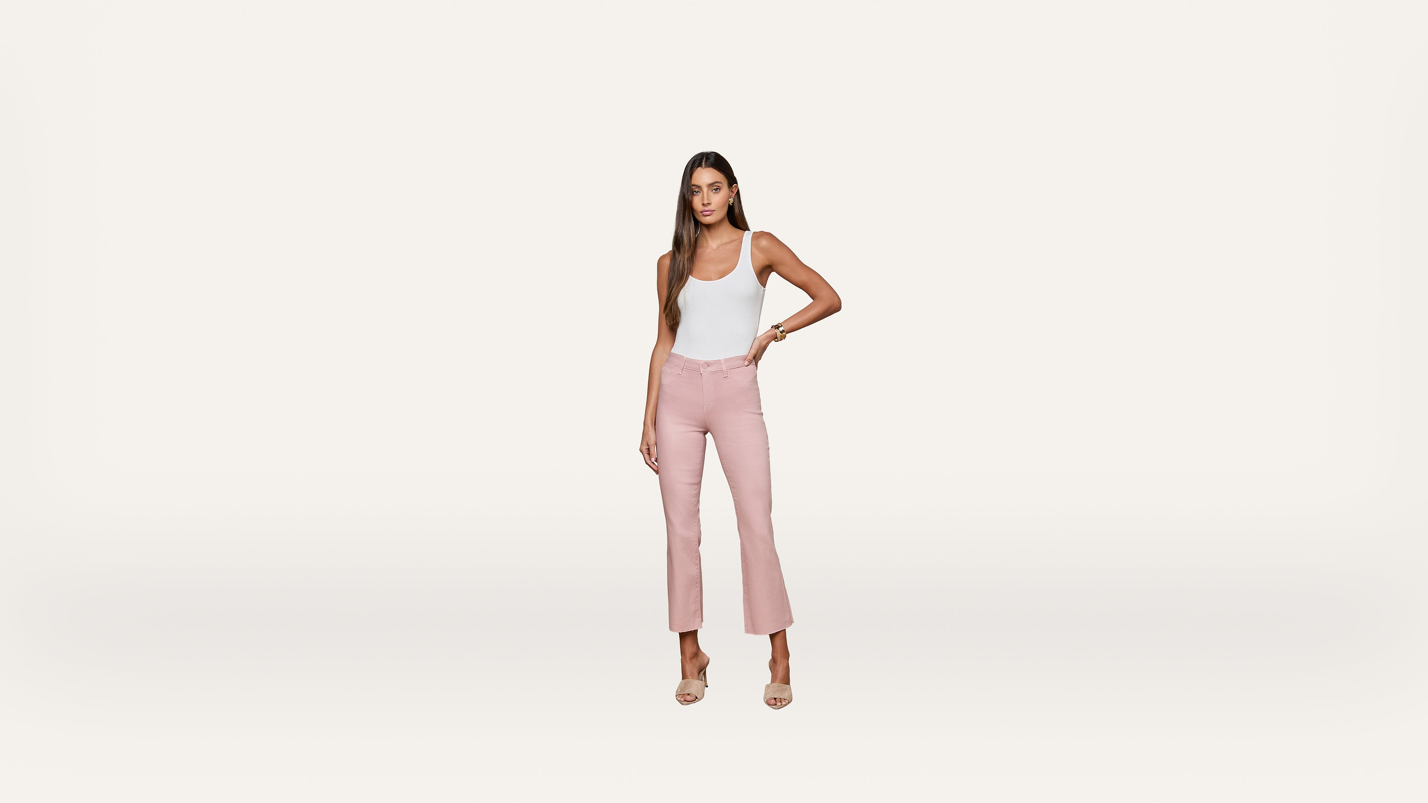 A woman stands against a plain white background wearing a white tank top, light pink high-waisted pants, and nude heels, with one hand on her hip.