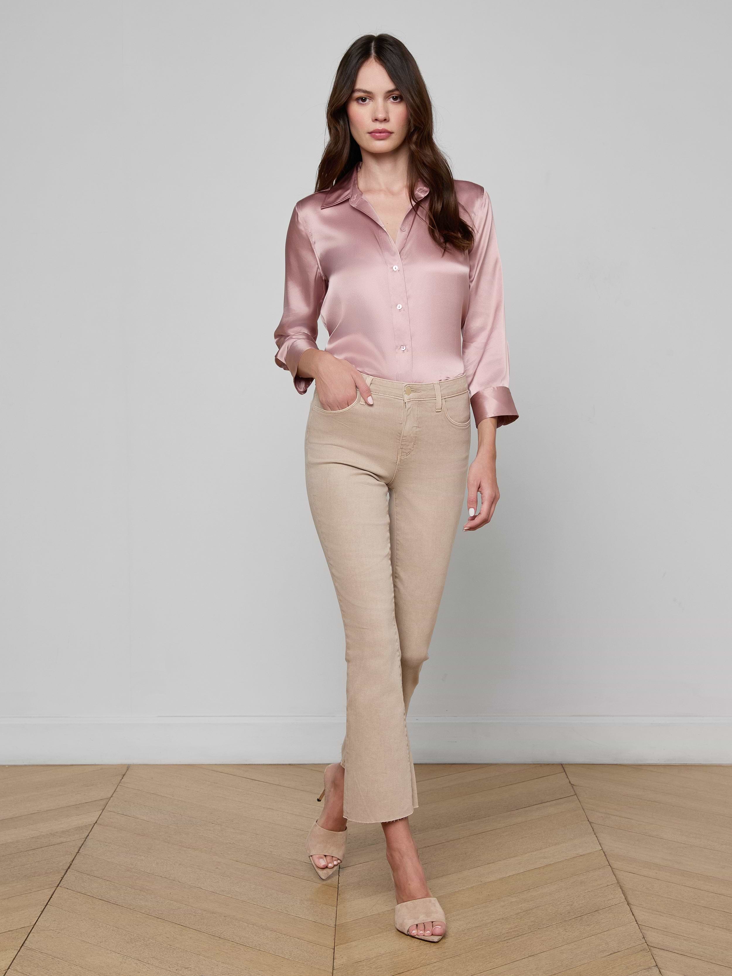 A woman stands against a white background wearing a silky pink blouse, the Kendra Cropped Flare Jean in neutral tan super stretch denim, and beige heeled sandals, with one hand in her pocket.