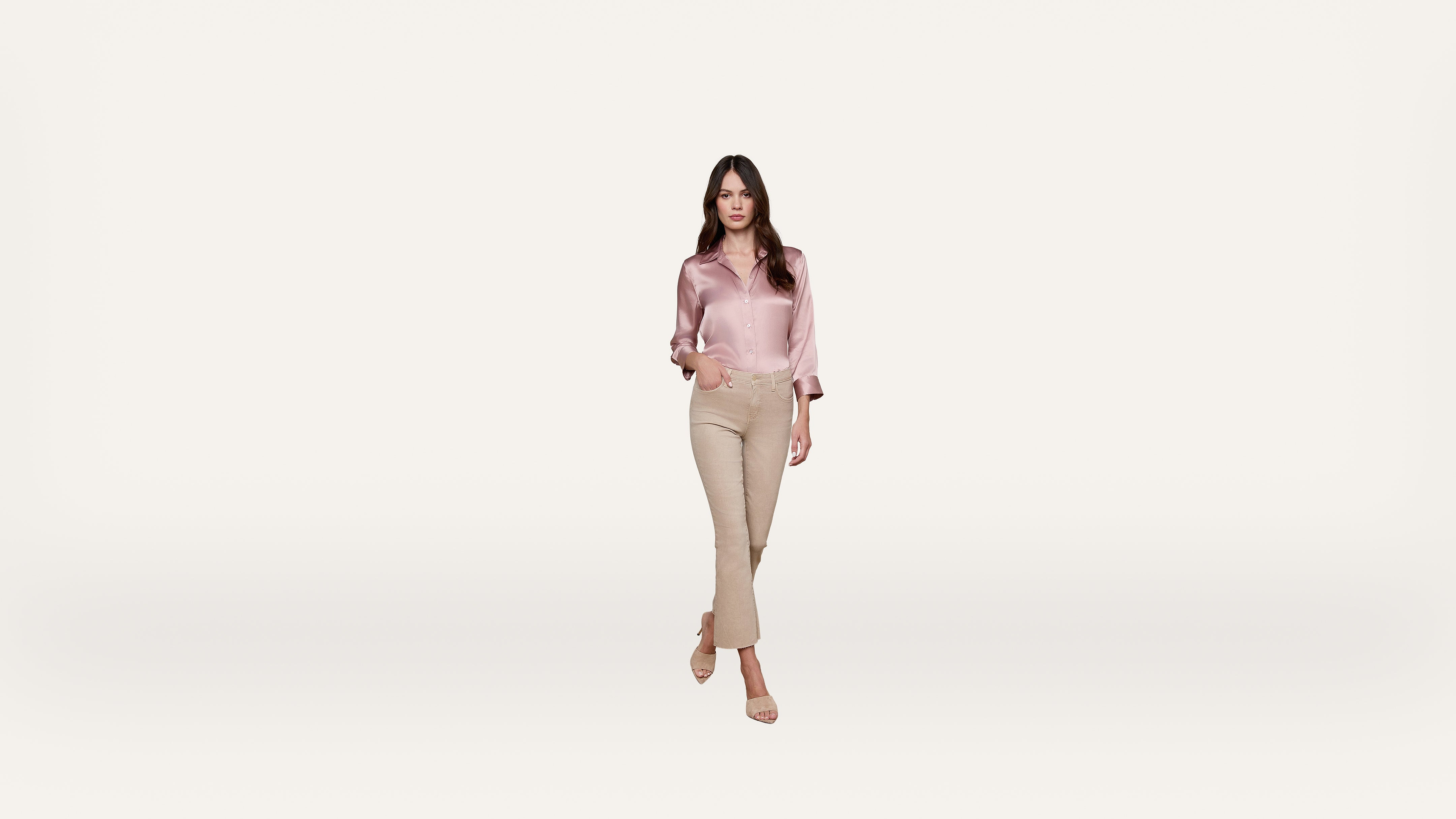 Woman with long brown hair wearing a pink satin blouse and beige pants walks forward against a plain white background.