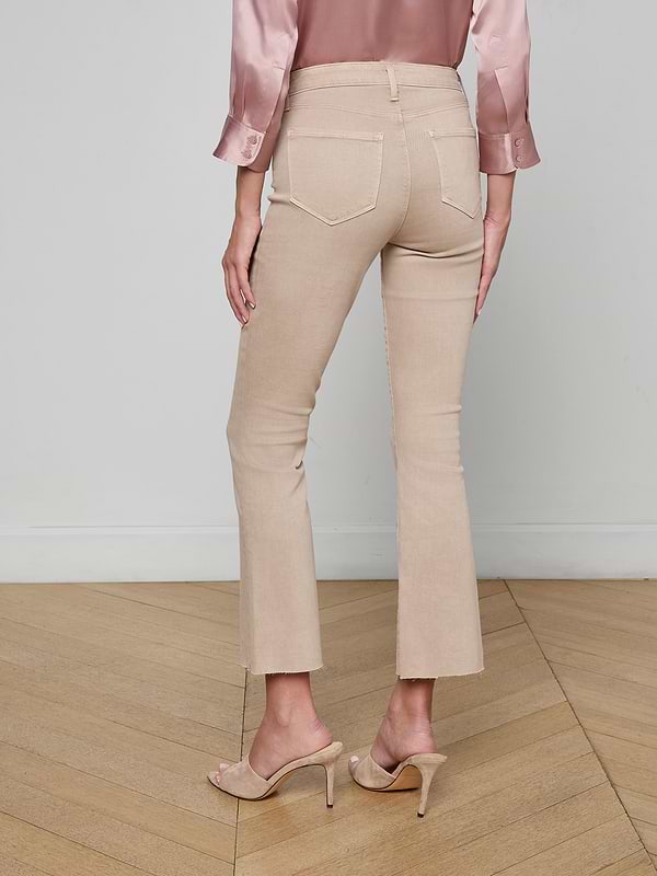 A woman, standing with her back to the camera against a plain background, is wearing Kendra Cropped Flare Jeans in a neutral tan shade, paired with a silky pink blouse and nude high-heeled sandals.