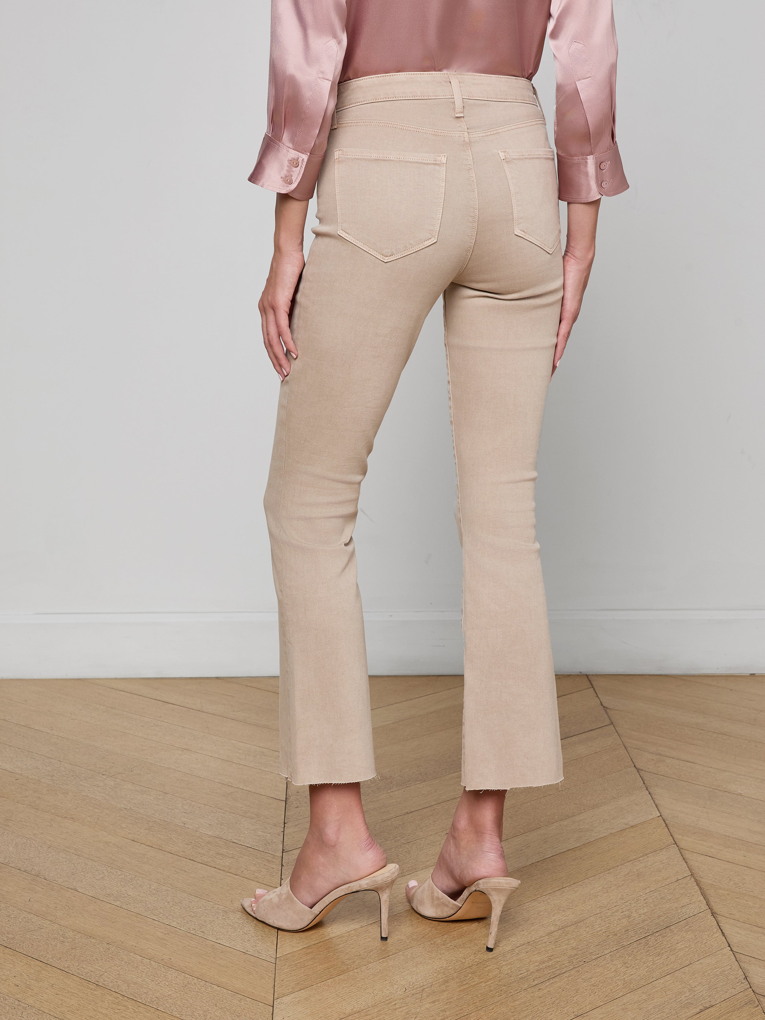 A woman, standing with her back to the camera against a plain background, is wearing Kendra Cropped Flare Jeans in a neutral tan shade, paired with a silky pink blouse and nude high-heeled sandals.