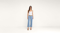 Woman wearing sunglasses, a white tank top, light blue wide-leg pants, a tan belt, and heels stands against a plain white background.