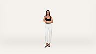 A woman stands with arms crossed, wearing a black top, white pants, and black heels, against a plain white background.