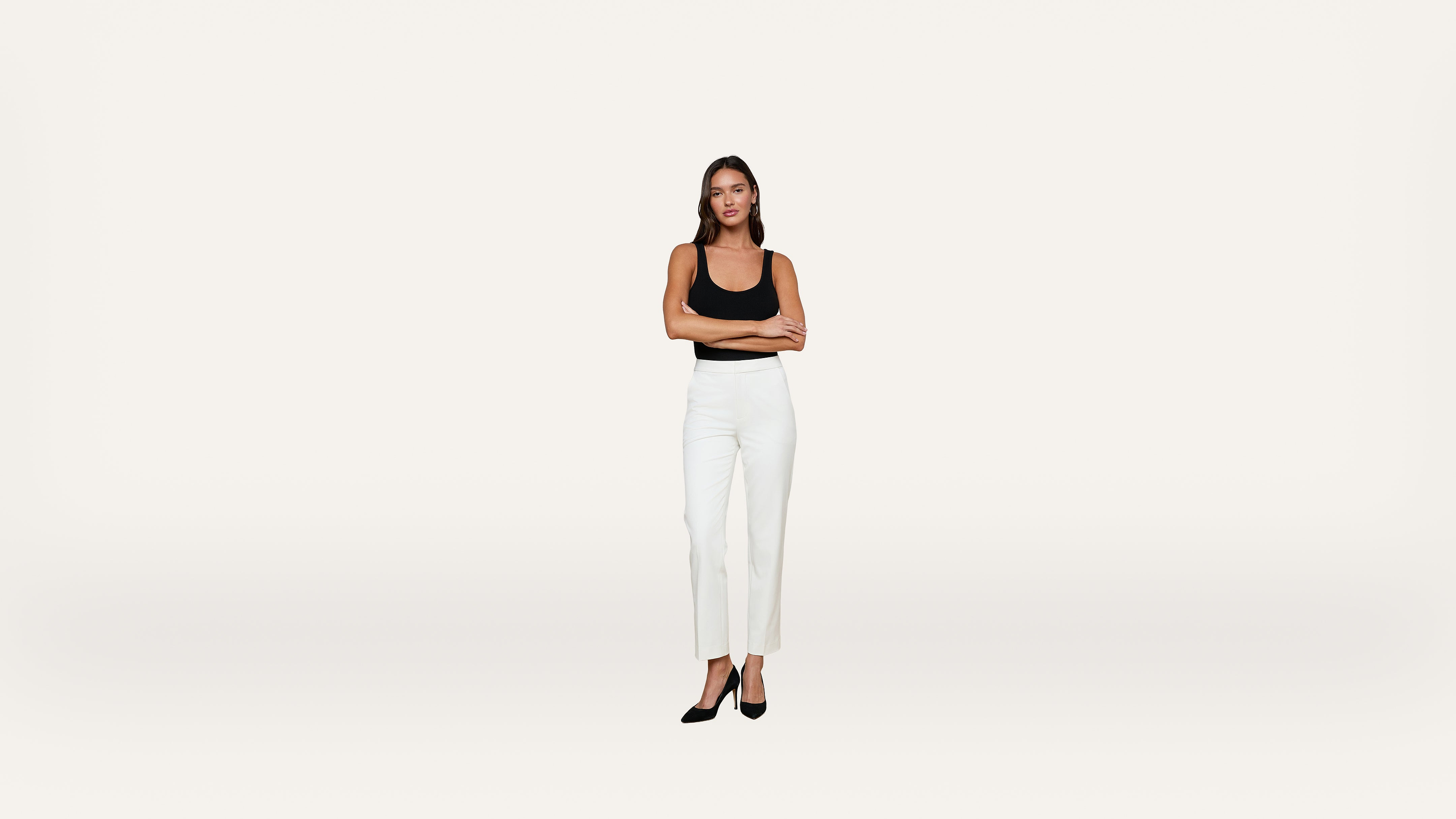 A woman stands with arms crossed, wearing a black top, white pants, and black heels, against a plain white background.