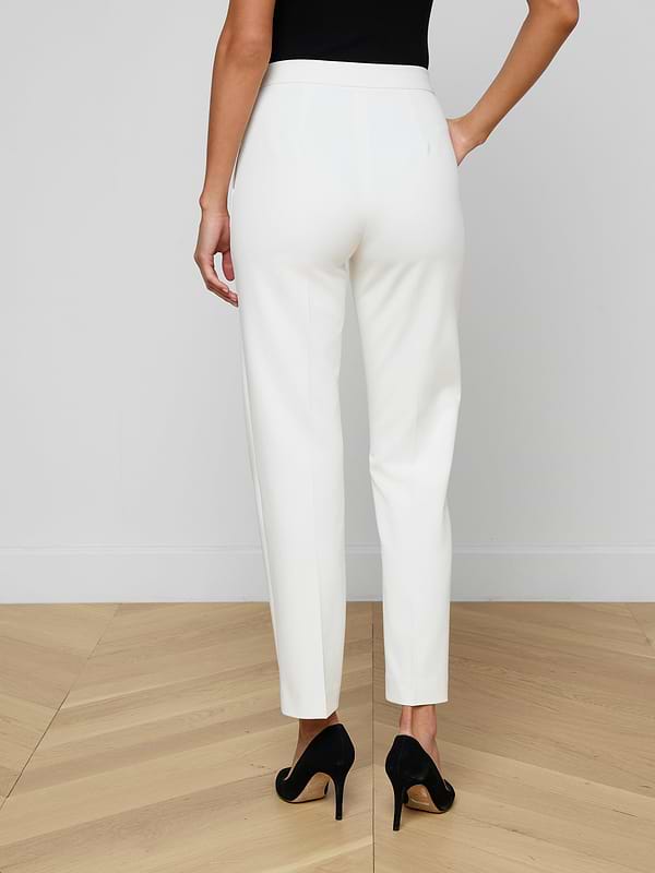 A woman stands facing away against a plain light background, showcasing the back view of the Rebel Trouser—white, high-rise straight-leg trousers crafted from stretch suiting fabric—and wearing black high heels.