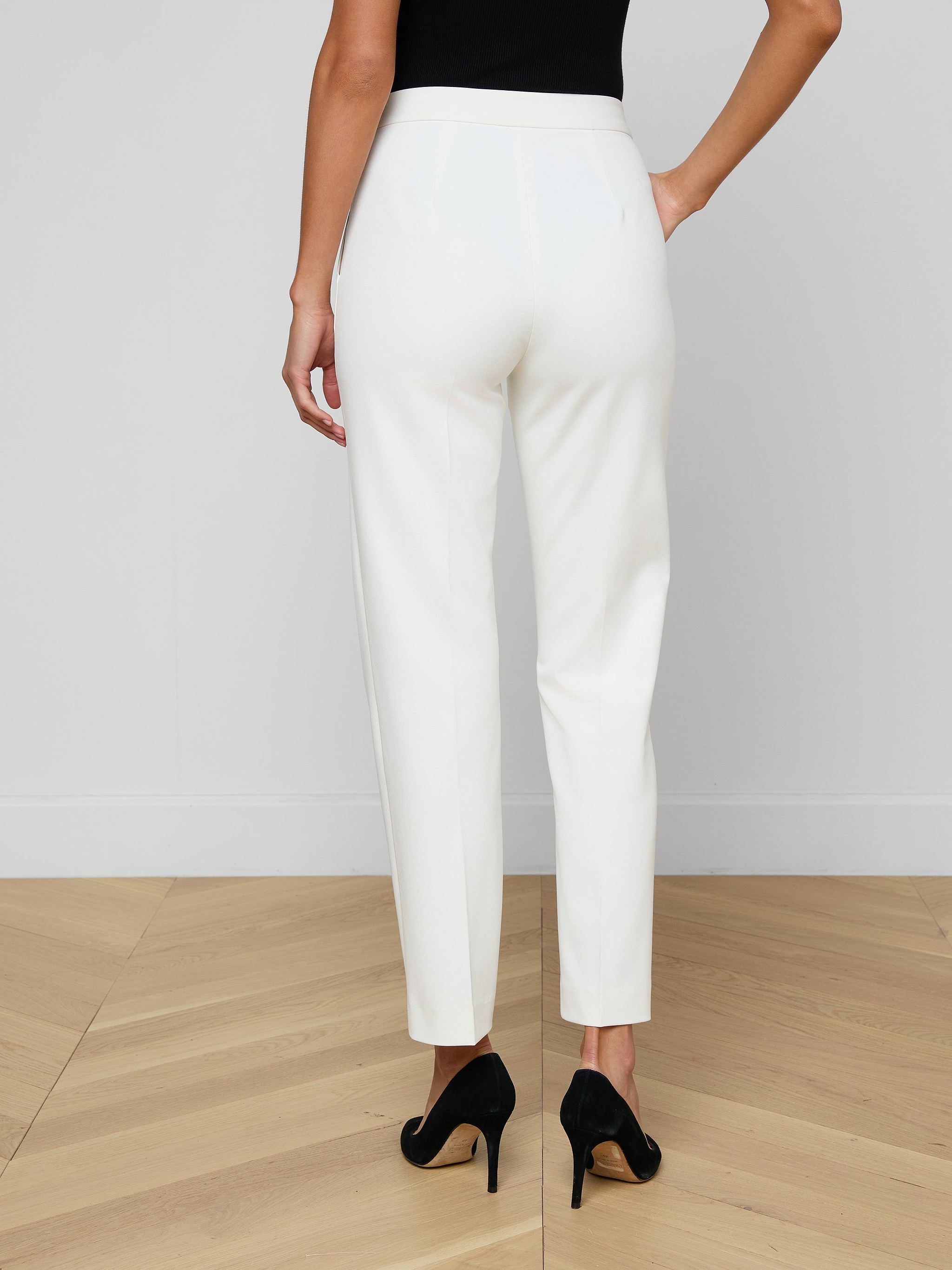 A woman stands facing away against a plain light background, showcasing the back view of the Rebel Trouser—white, high-rise straight-leg trousers crafted from stretch suiting fabric—and wearing black high heels.
