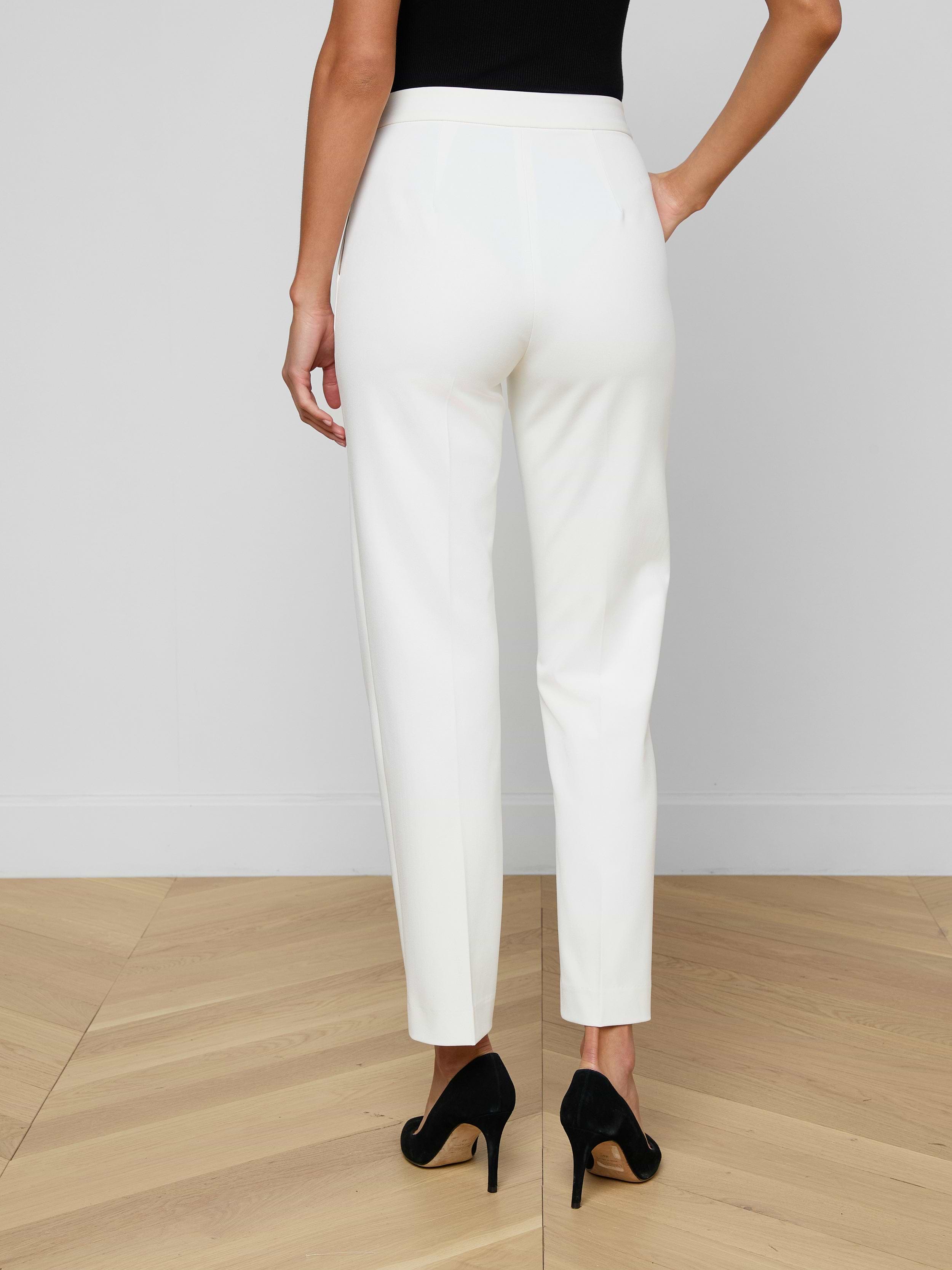 A woman stands facing away against a plain light background, showcasing the back view of the Rebel Trouser—white, high-rise straight-leg trousers crafted from stretch suiting fabric—and wearing black high heels.