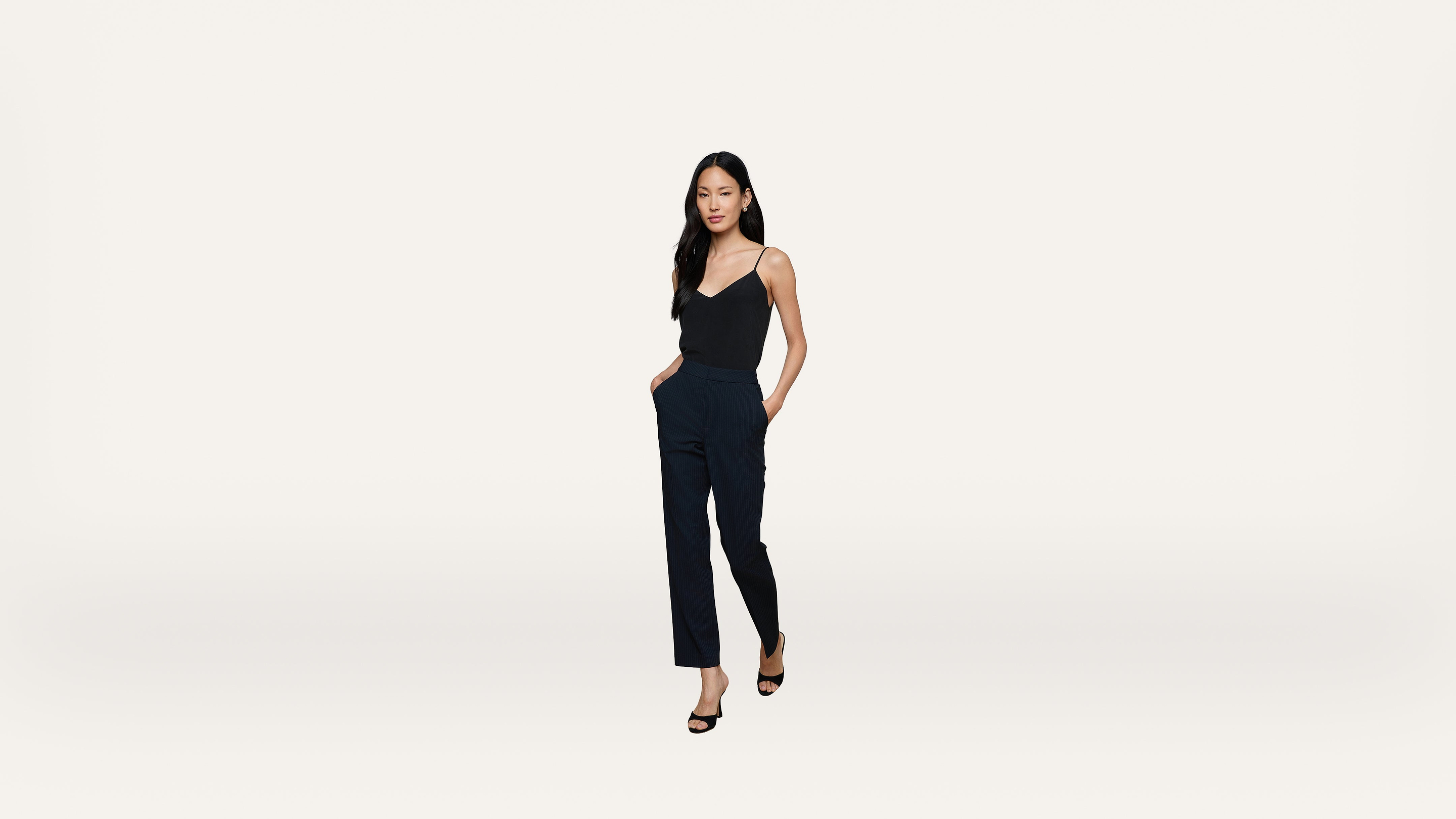 A woman with long black hair wears a black sleeveless top, navy blue pants, and black heels, standing with hands in pockets against a plain white background.