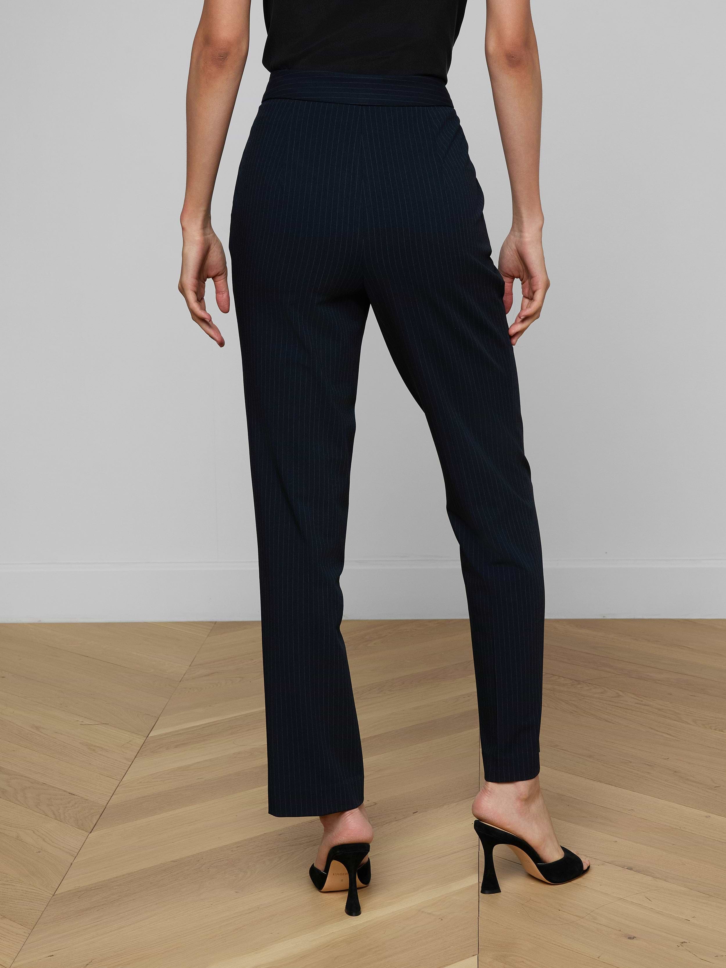 A person wearing the Rebel Pinstriped Trouser, featuring a high-rise, straight-leg silhouette with navy pinstripes, and black heeled shoes stands with their back to the camera against a plain light background.