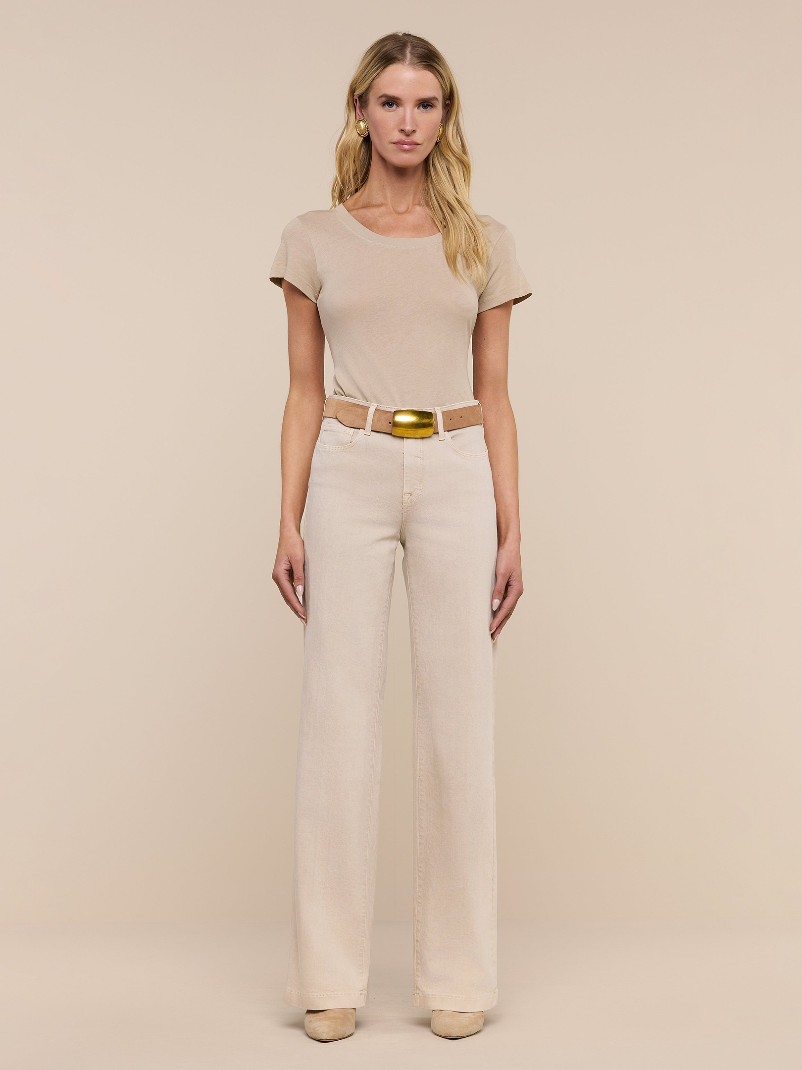 A woman stands facing forward, wearing a beige short-sleeve top, beige shoes, and the Clayton Wide-Leg Jean in a matching high-rise, tall wide-leg style with a belt, against a plain white background.