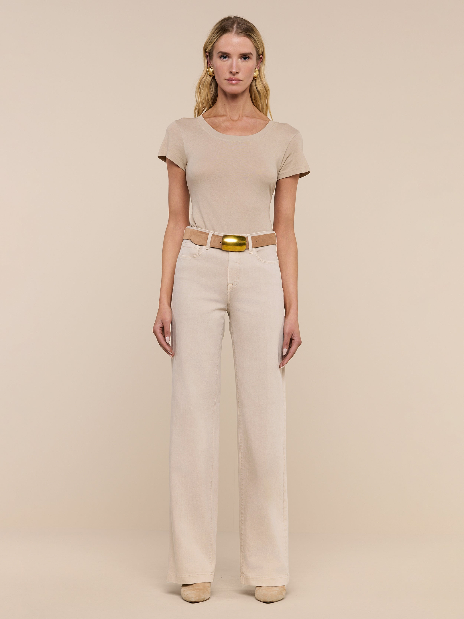 A woman stands facing forward in a beige short-sleeve top, the Clayton Wide-Leg Jean in stretch denim with a belt, and beige shoes, against a plain white background.