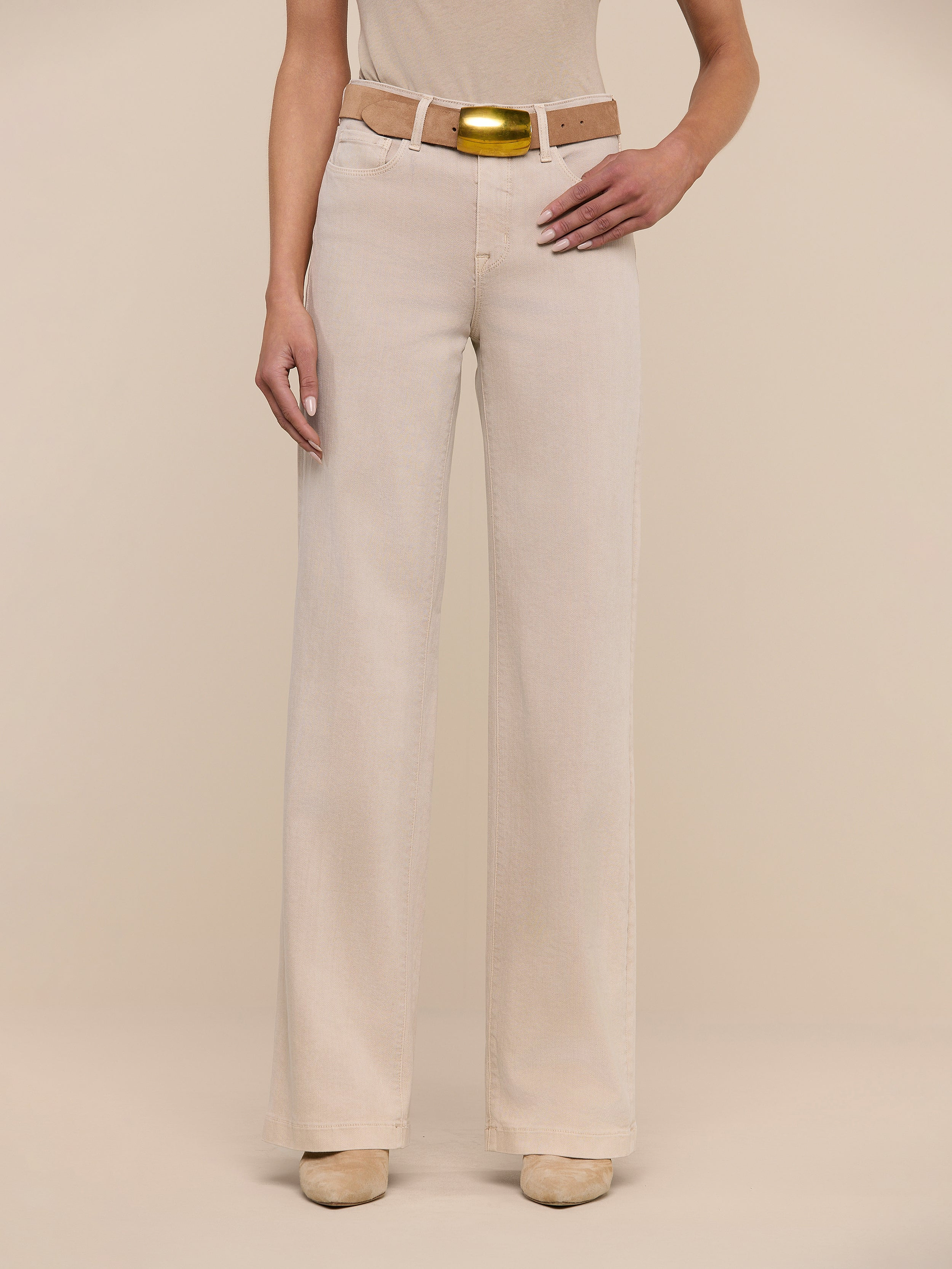 A person wearing the Clayton Wide-Leg Jean in beige, paired with a matching beige shirt and a tan belt with a gold buckle, stands against a plain white background. Their head is not visible in the image.