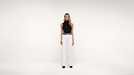 A woman stands facing forward against a plain white background, wearing a sleeveless black turtleneck top, white wide-leg trousers, a white belt with a gold buckle, and black shoes.