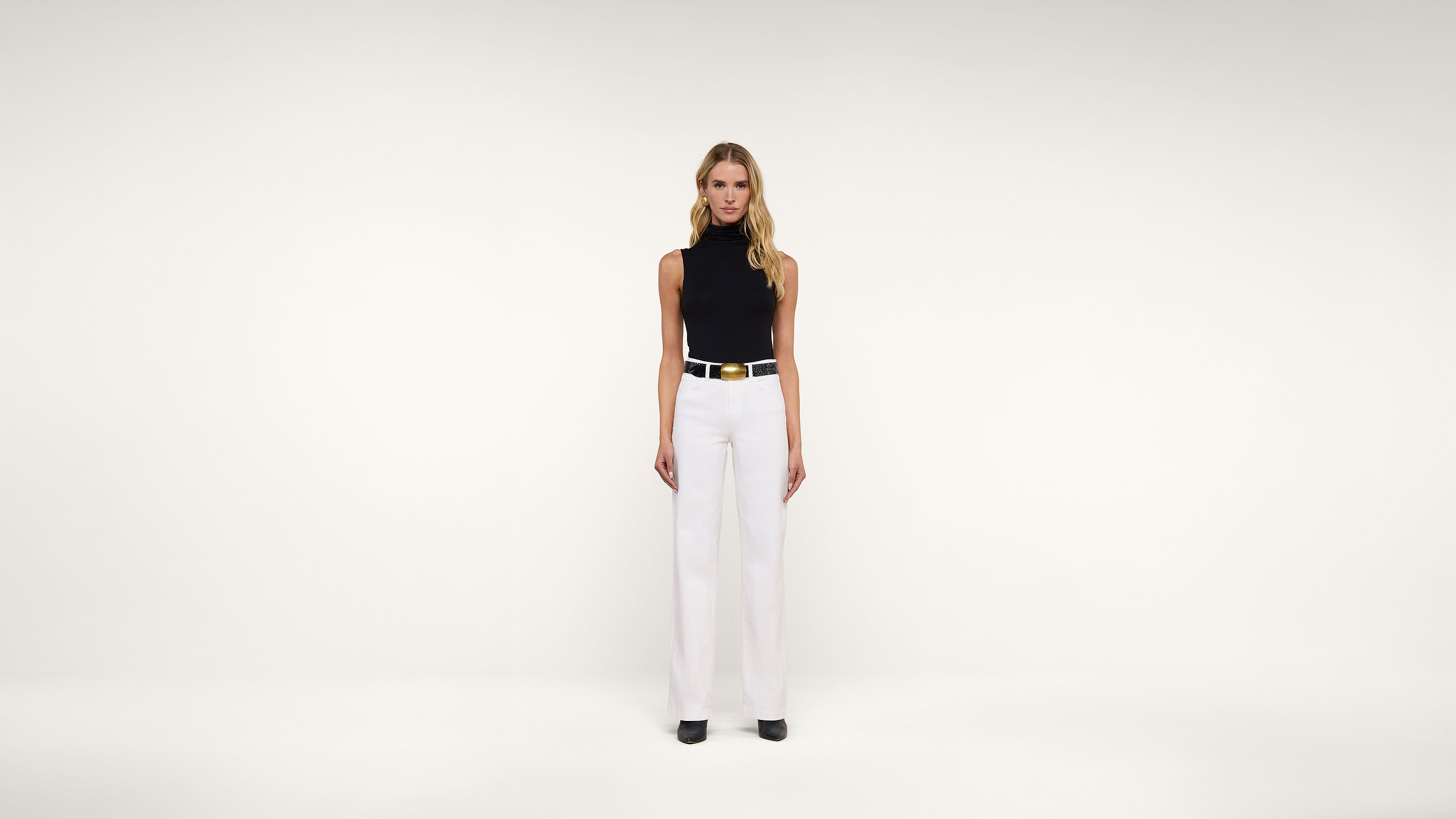 A woman stands facing forward against a plain white background, wearing a sleeveless black turtleneck top, white wide-leg trousers, a white belt with a gold buckle, and black shoes.