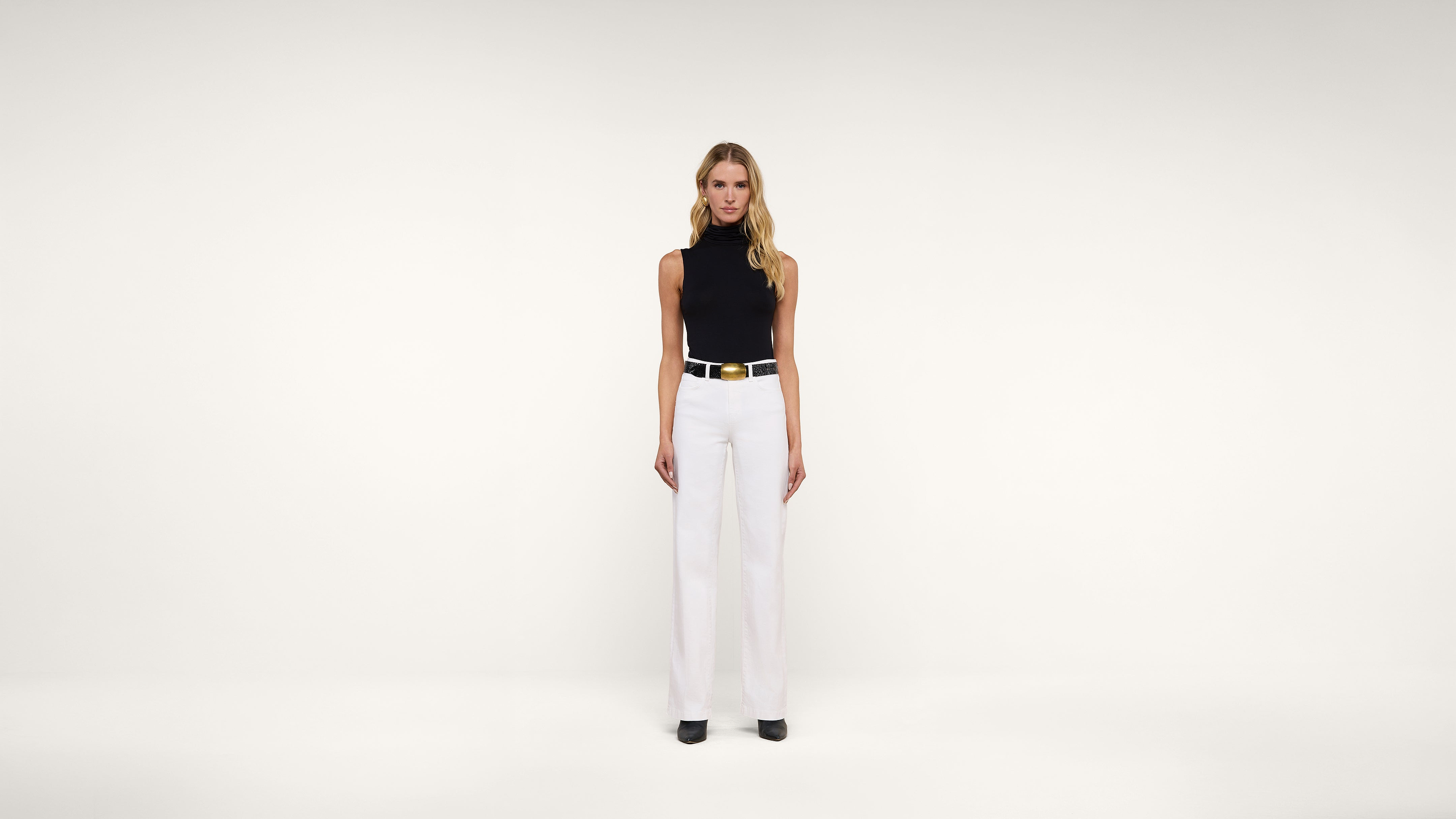 A woman stands facing forward against a plain white background, wearing a sleeveless black turtleneck top, white wide-leg trousers, a white belt with a gold buckle, and black shoes.