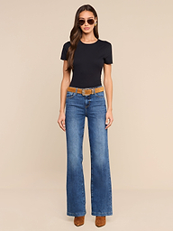 A woman wearing sunglasses, a black t-shirt, the Clayton Wide-Leg Jean in blue stretch denim, a brown belt, and brown shoes stands against a plain white background.
