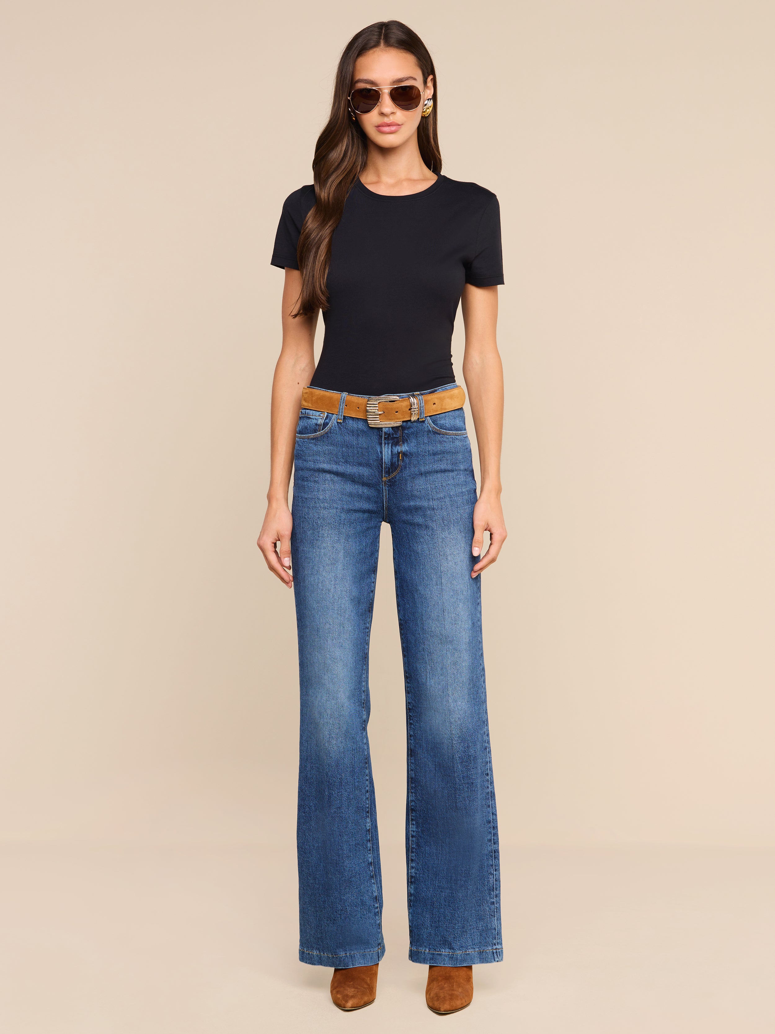 A woman wearing sunglasses, a black t-shirt, the Clayton Wide-Leg Jean in blue stretch denim, a brown belt, and brown shoes stands against a plain white background.